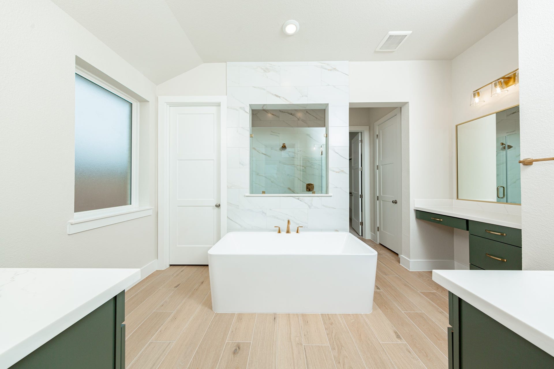 This elegant primary bathroom features a stylish dual-vanity layout with stained wood cabinetry, sleek brushed gold fixtures, and clean-lined framed mirrors. Bright quartz countertops and modern lighting create a fresh, upscale feel, while the space opens seamlessly to the primary suite beyond, enhancing both flow and functionality.