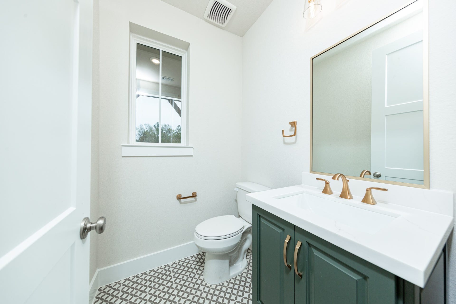 The first-floor powder room offers convenience and a modern aesthetic. It features a stained cabinet, quartz countertop, matte gold sleek faucet and a matching wall-mounted towel ring, window, tile flooring, and a framed mirror.