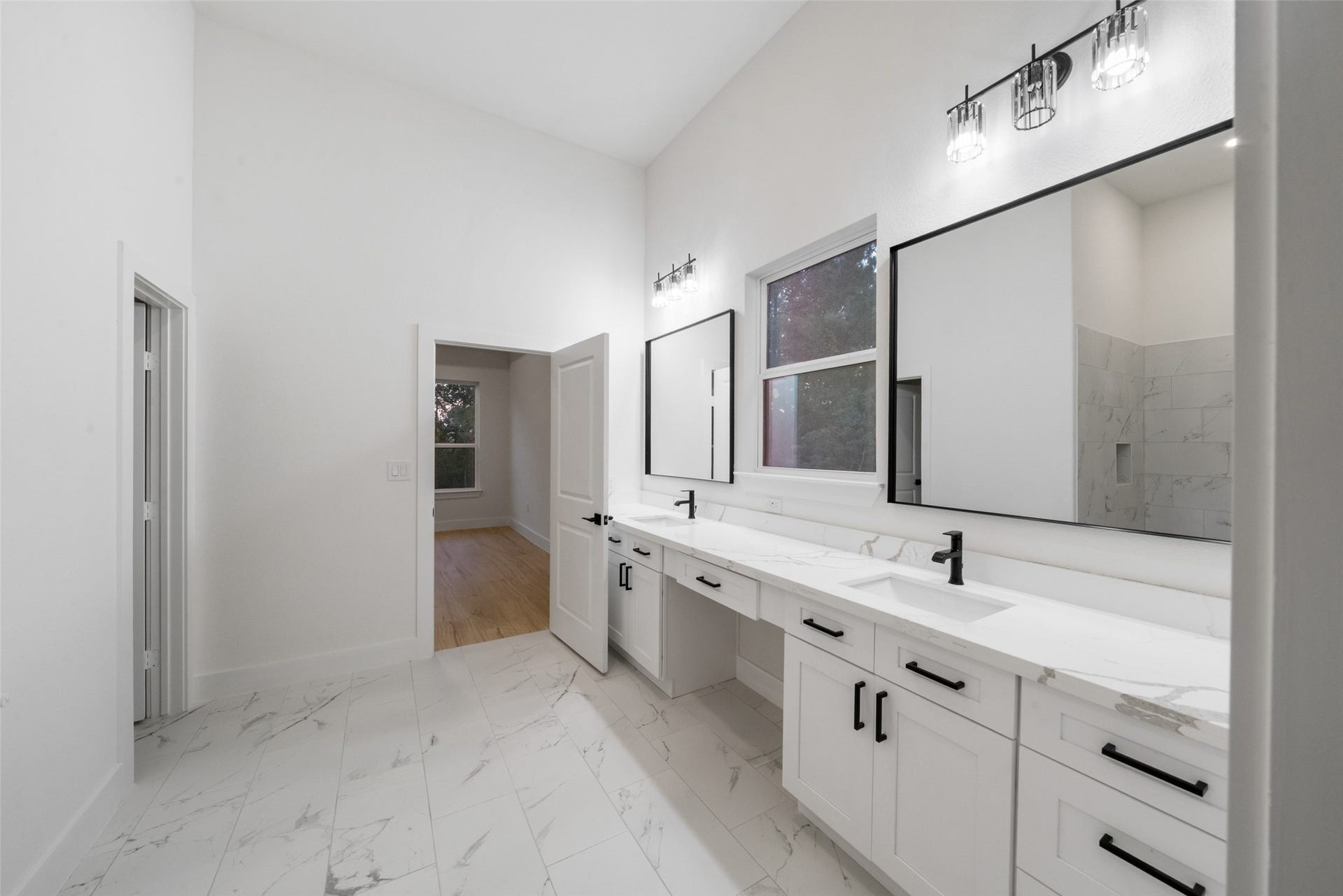 4833 W Fork Boulevard, Conroe, TX 77304 Luxurious primary bathroom showcasing an extended dual-sink vanity with elegant countertops, individual framed mirrors, and sophisticated black fixtures. A large window welcomes natural light, beautifully complementing the room's bright, timeless finishes.