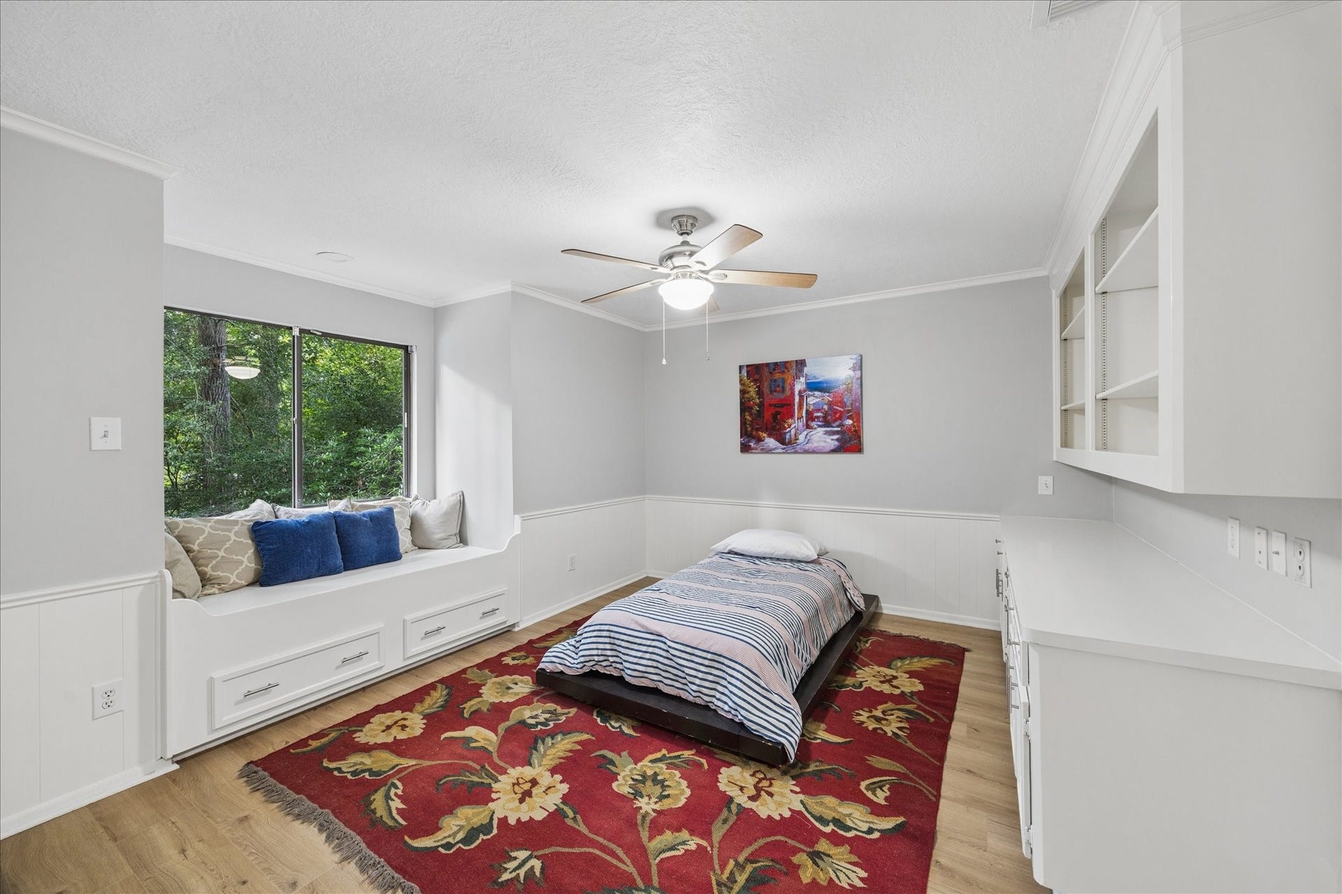2411 Oakbank Drive, Kingwood, TX 77339 The 2nd-floor bedroom features a built-in window seat with storage, a ceiling fan, and a comfortable bed. The neutral walls and wood flooring create a warm atmosphere, complemented by a vibrant floral rug. Built-in shelves and a desk area offer ample storage and workspace.