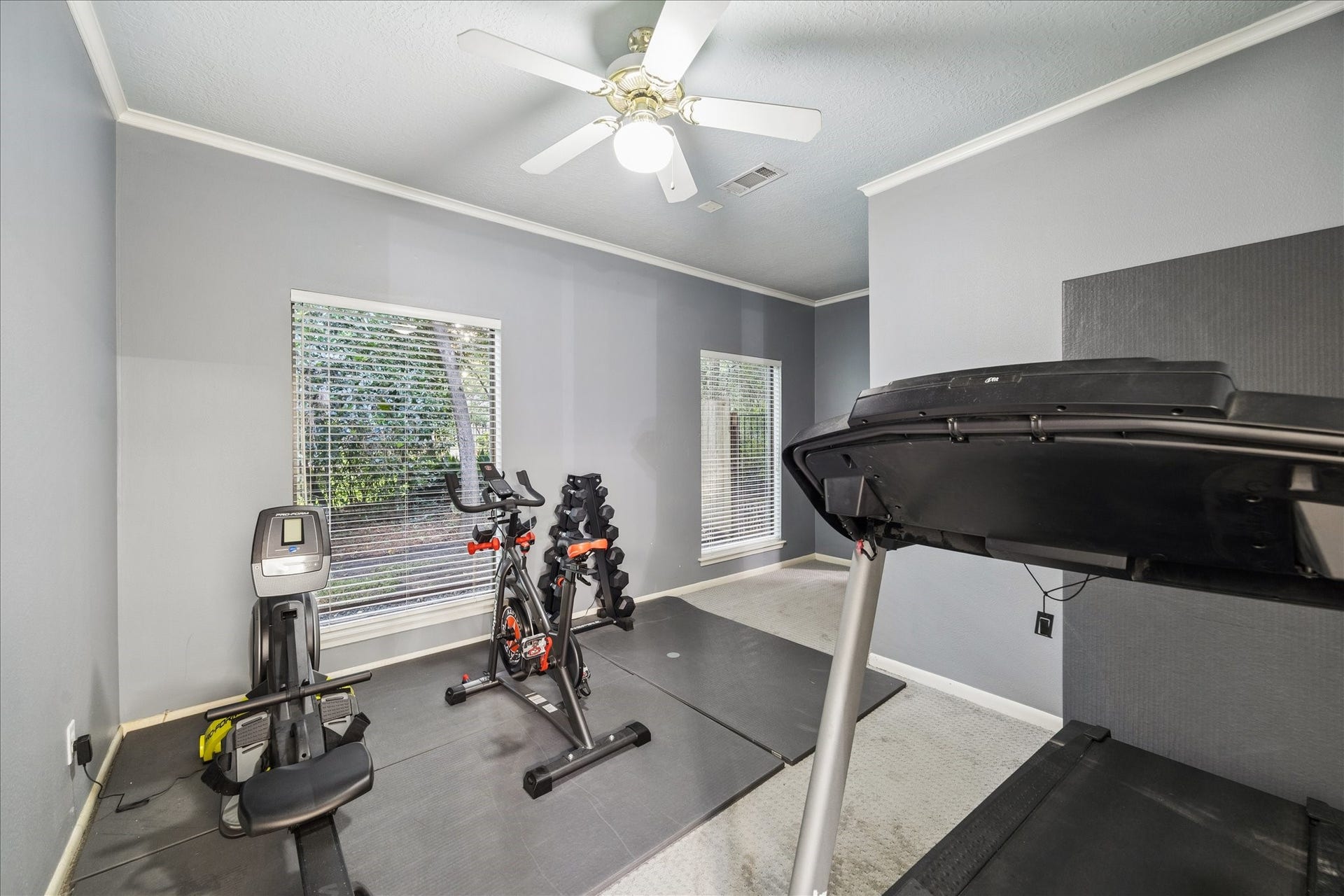 2411 Oakbank Drive, Kingwood, TX 77339 Next to the primary suite is a home gym with a treadmill, exercise bike, and rowing machine, featuring two large windows for natural light and a ceiling fan for ventilation.