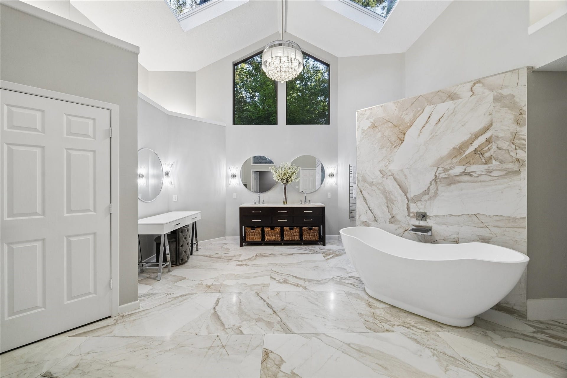 2411 Oakbank Drive, Kingwood, TX 77339 This luxurious primary bathroom features elegant marble flooring and walls, a freestanding bathtub, and a large window that fills the space with natural light. The room is complemented by a modern vanity with double sinks, two round mirrors, and a chandelier for added sophistication.