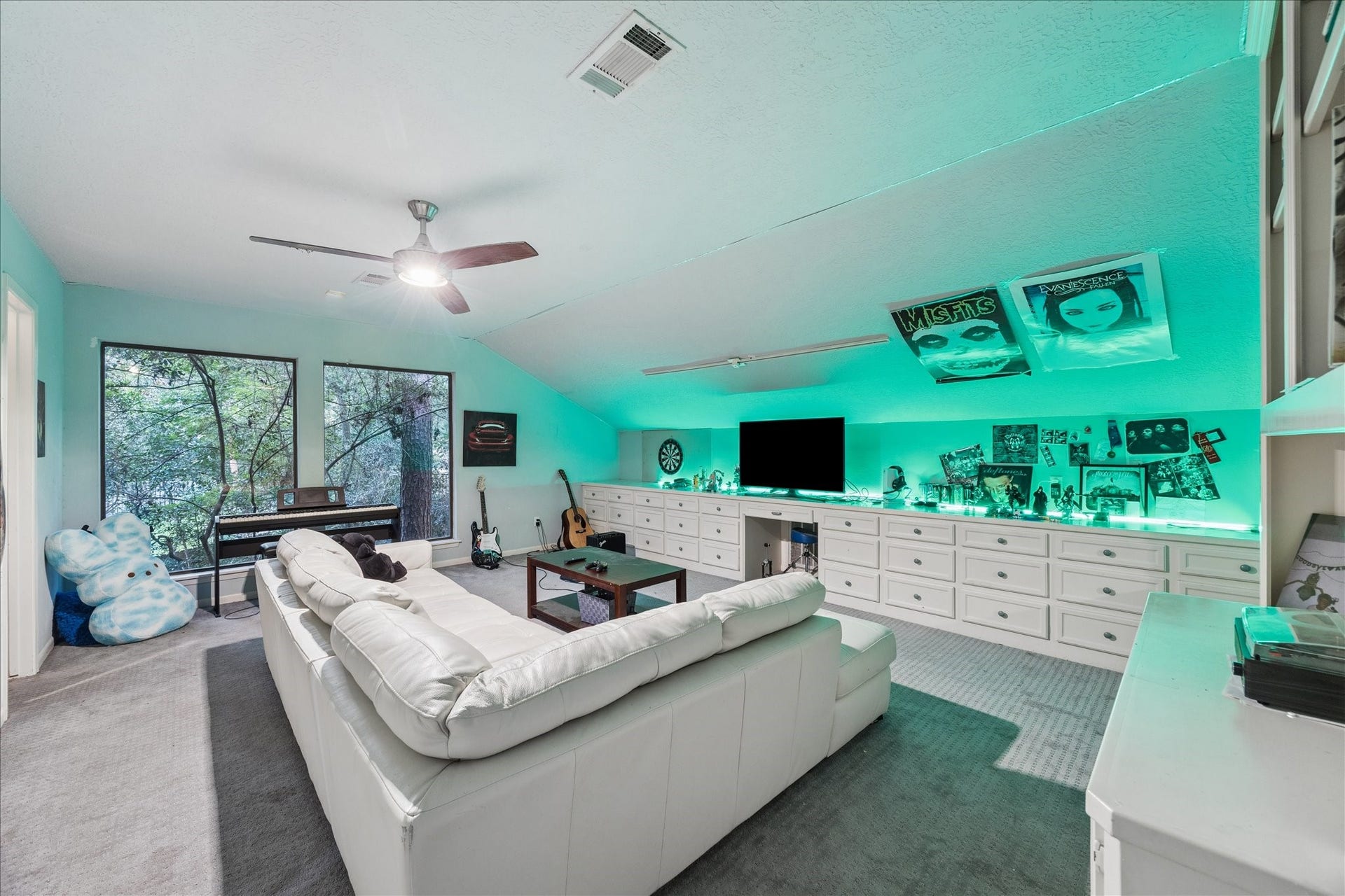 2411 Oakbank Drive, Kingwood, TX 77339 This 2nd-floor spacious entertainment room features a cozy white sectional, ambient lighting, and a wall-mounted TV. It includes ample storage with built-in cabinets and large windows providing natural light and views of the backyard.