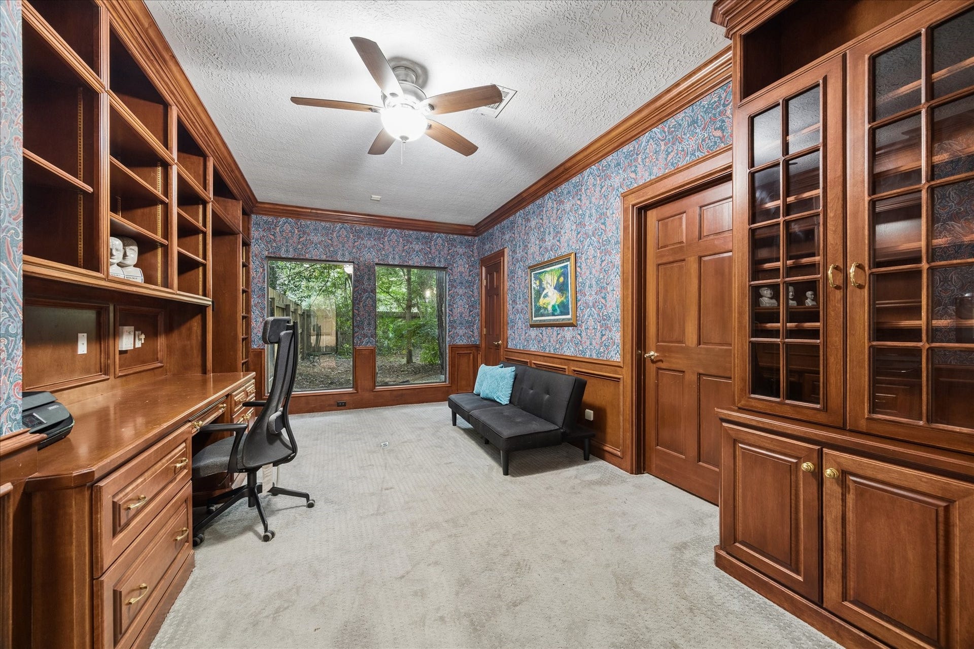 2411 Oakbank Drive, Kingwood, TX 77339 This is the 2nd home office next to the primary suite, with rich wood built-in shelves and cabinets. It features a large desk area, a comfortable chair, and a sofa. Two large windows provide natural light and a view of greenery, while a ceiling fan adds comfort. The room has a classic, elegant feel with its wood details and patterned wallpaper.