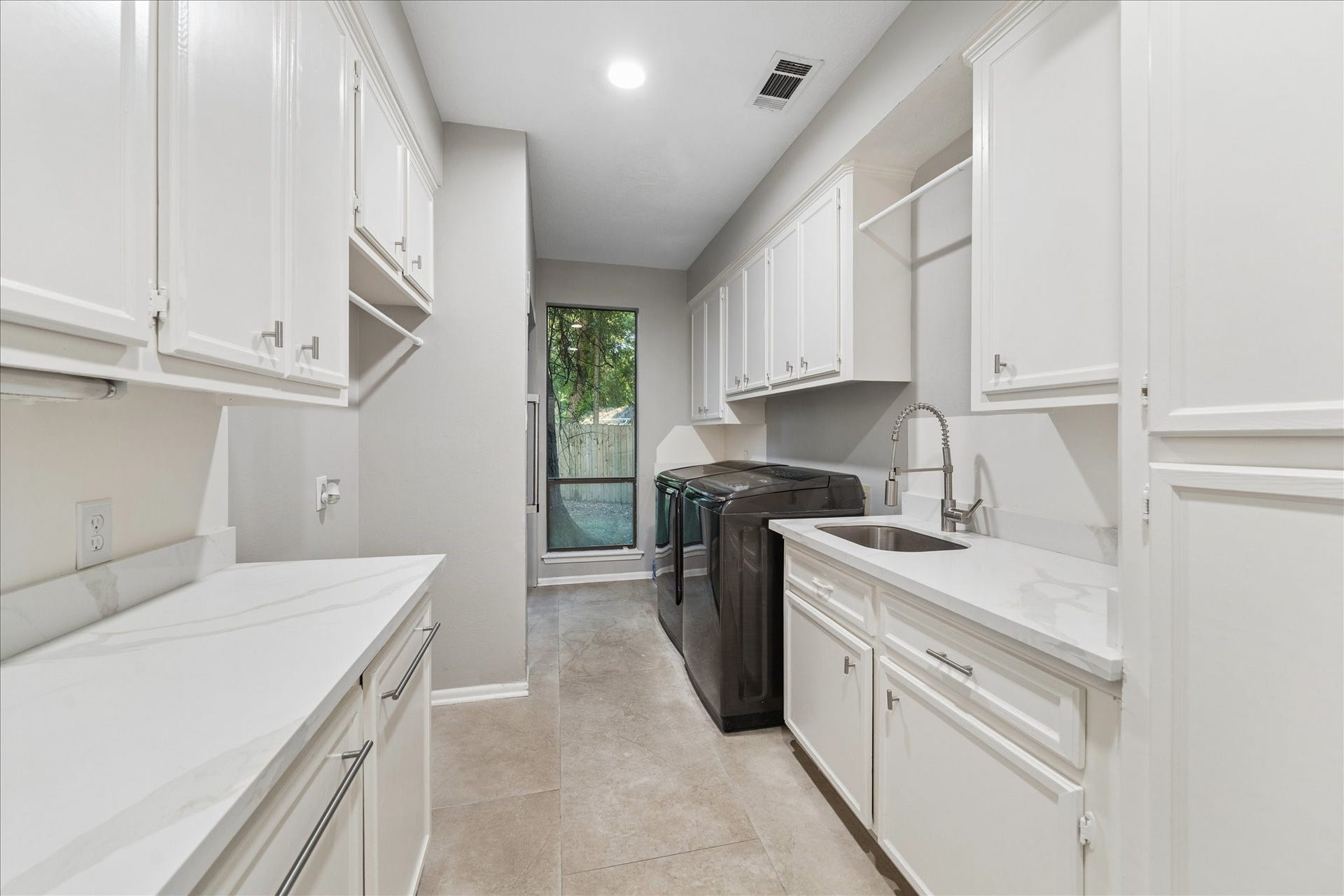 2411 Oakbank Drive, Kingwood, TX 77339 The laundry room features sleek white cabinetry, ample storage, and counter space. It features a utility sink and a window providing natural light. The neutral tile flooring complements the clean, bright aesthetic.