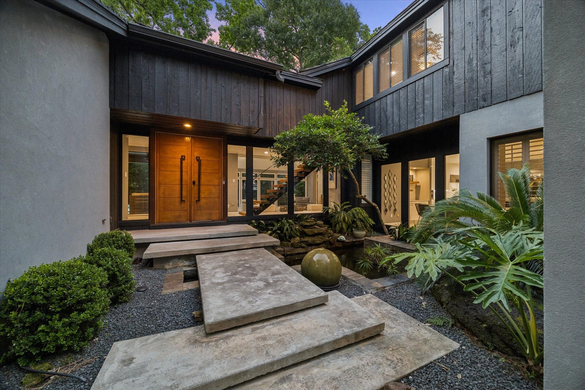 2411 Oakbank Drive, Kingwood, TX 77339 Stunning entrance featuring a minimalist stone pathway over a serene water feature and koi pond framed by lush greenery and sleek, dark wood paneling. Large windows provide a glimpse into the inviting interior, creating a harmonious blend of nature and architecture.