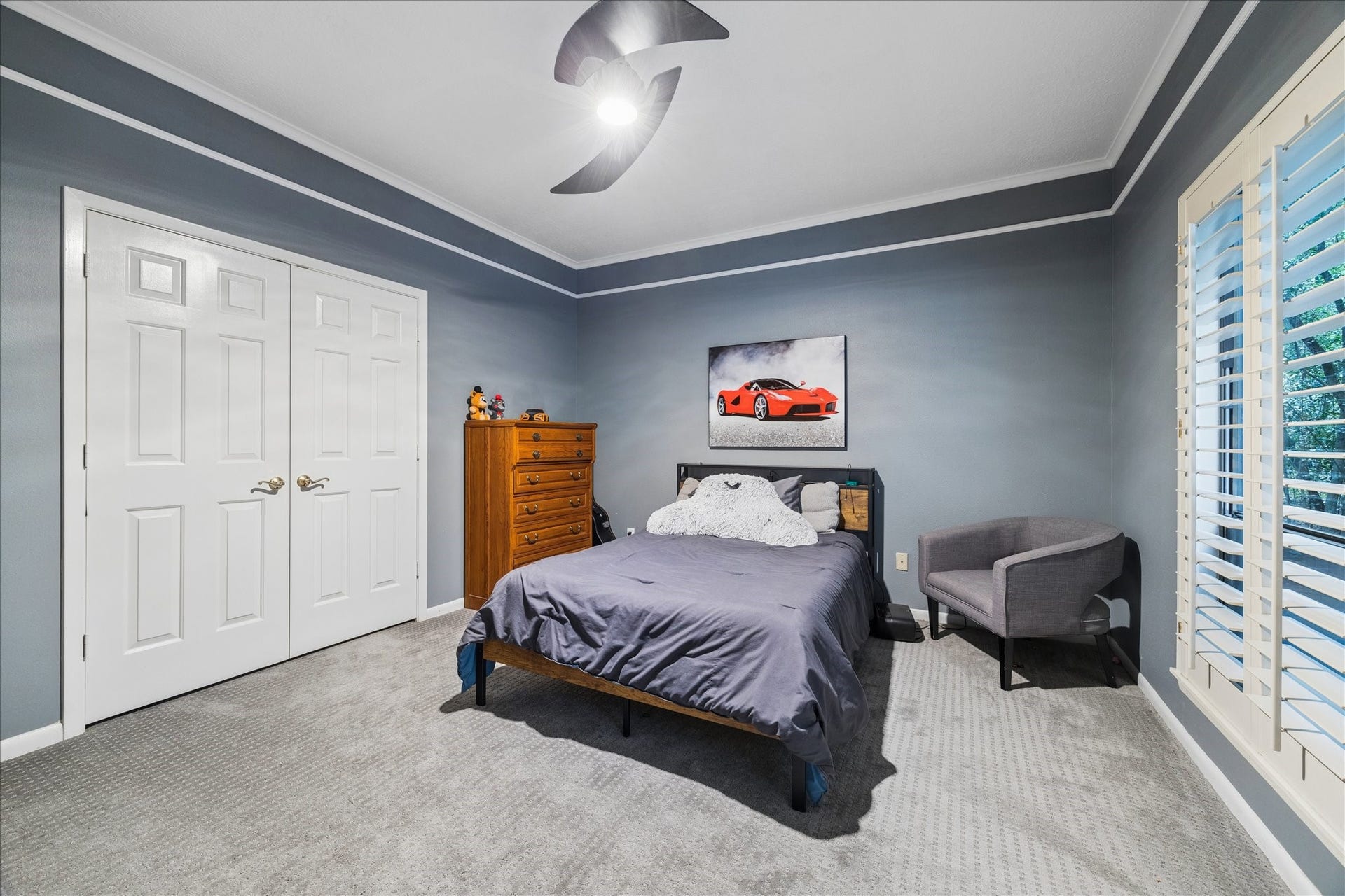 2411 Oakbank Drive, Kingwood, TX 77339 The first-floor guest bedroom features a cozy atmosphere with gray walls, a carpeted floor, and a ceiling fan. Natural light enters through large shuttered windows, and there's a closet with double doors for ample storage.