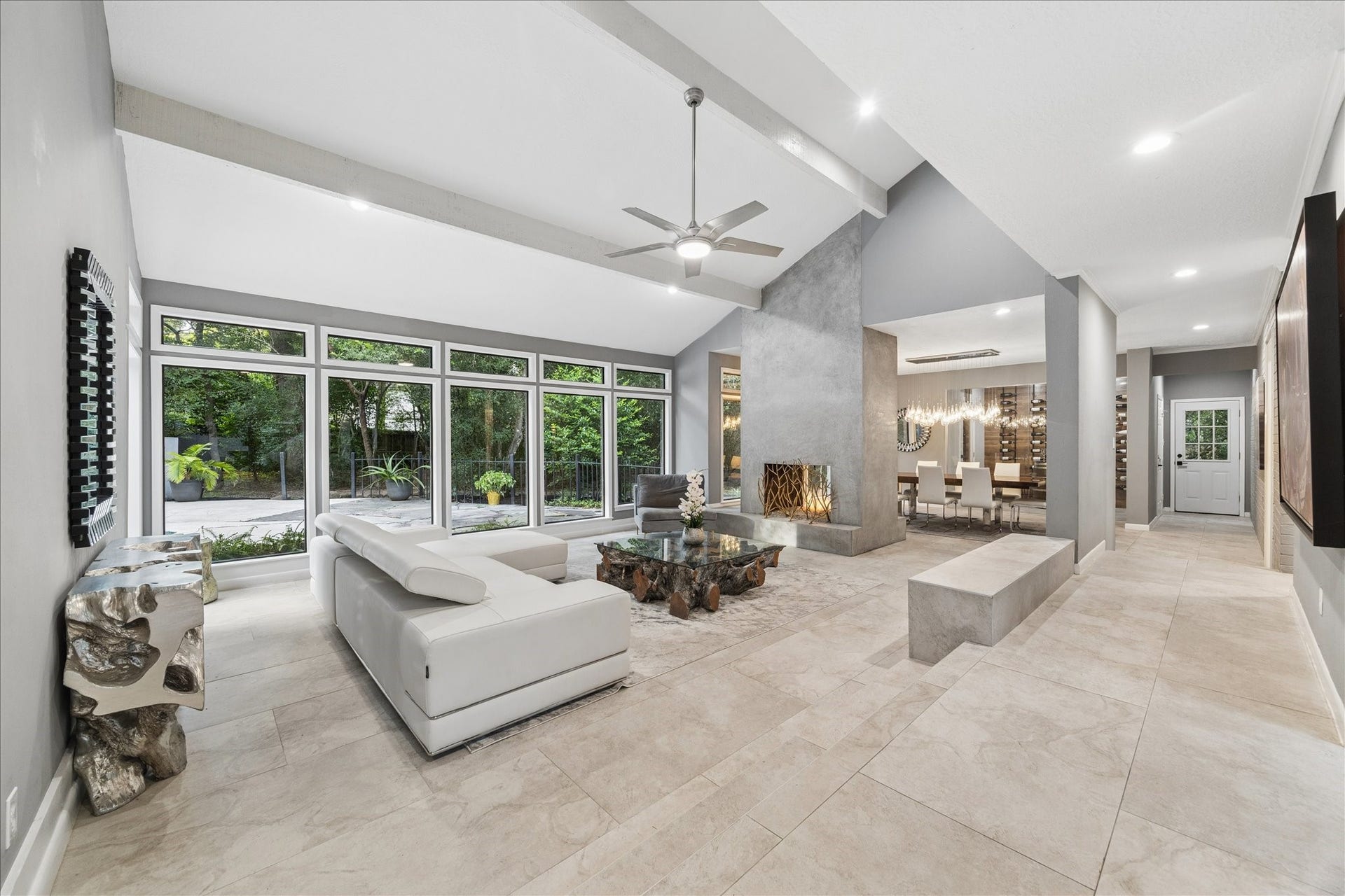 2411 Oakbank Drive, Kingwood, TX 77339 This modern living room features a sleek design with large floor-to-ceiling windows offering ample natural light and a view of lush greenery. It includes a contemporary fireplace, elegant dining area, and neutral-toned flooring, creating a spacious and inviting atmosphere.