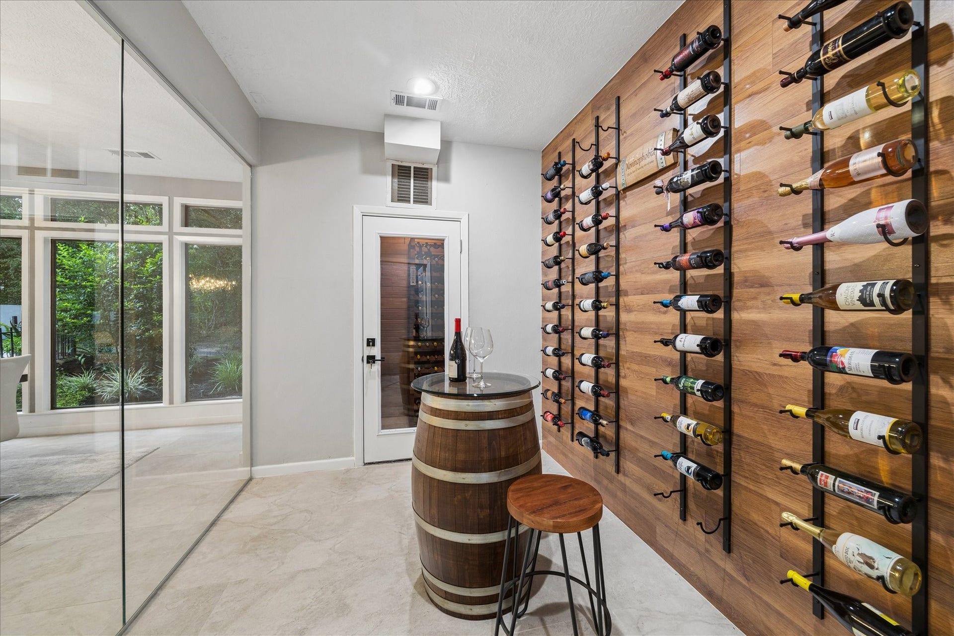 2411 Oakbank Drive, Kingwood, TX 77339 Enjoy your very own wine cellar and climate-controlled wine room. With a stylish wooden wall-mounted wine rack, a glass door, and a cozy tasting area featuring a barrel table and stool. Large windows provide a view of lush greenery, adding a serene backdrop.