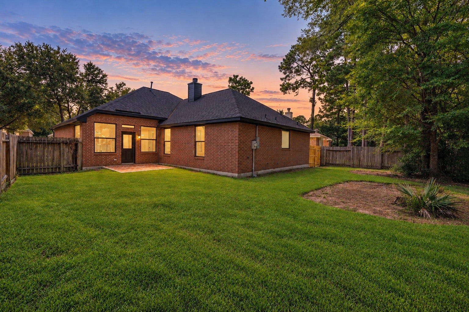 18 Griffin Hill Court, The Woodlands, TX 77382 A stunning twilight view of the rear exterior of 18 Griffin Hill Court reveals the home's beautiful brick construction bathed in warm orange and pink sunset hues. Mature trees create a natural privacy canopy around the backyard, while the glow of the evening sky sets a peaceful and magical scene for this remarkable home. (PHOTO MAY BE AI ENHANCED.)