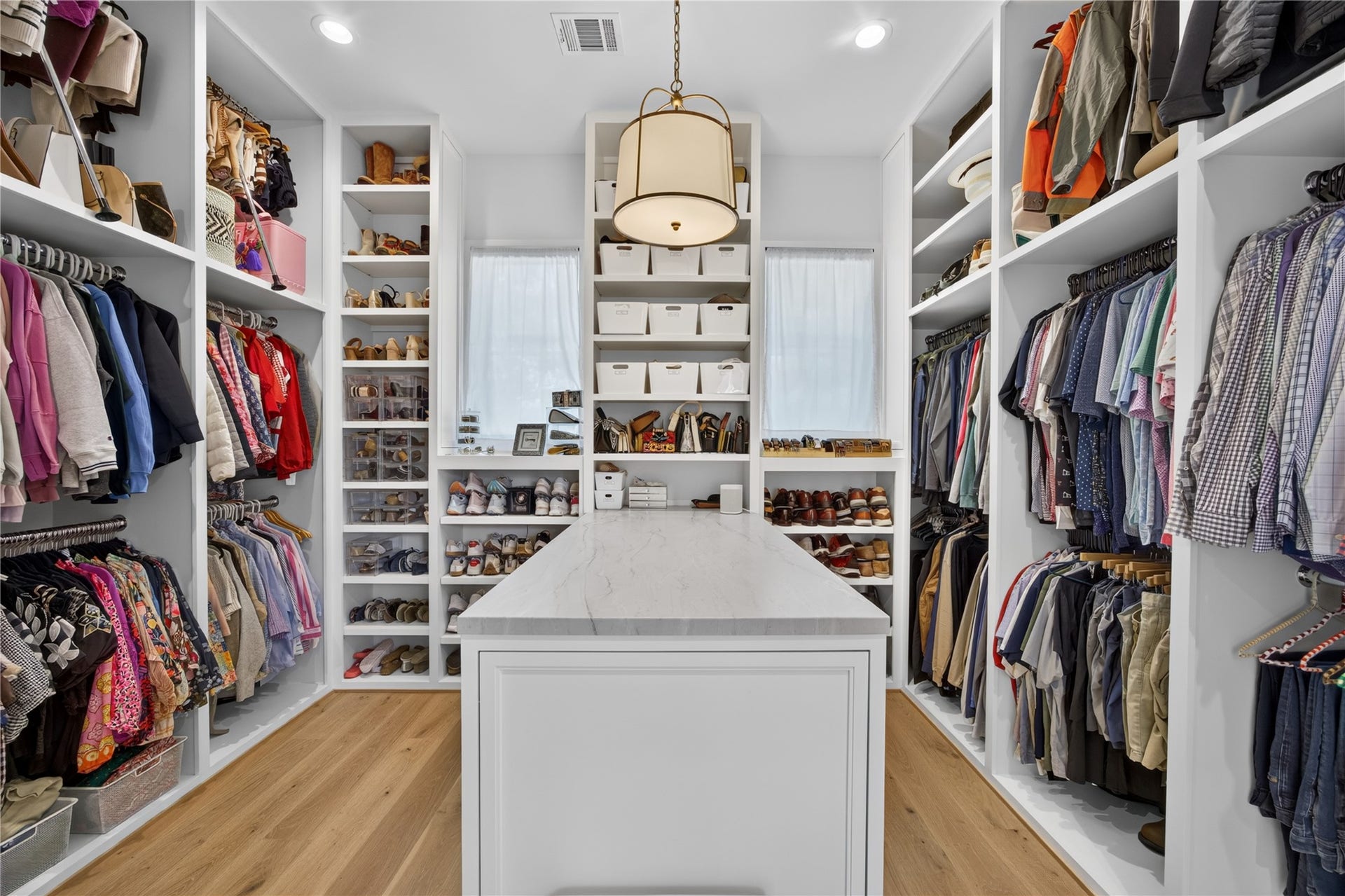 13915 Queensbury Lane, Houston, TX 77079 An exceptional walk-in closet designed for organization and convenience, featuring extensive custom built-in shelving, double hanging rods, dedicated shoe storage, and a spacious center island. Beautiful wood flooring, natural light from two windows, and elegant pendant lighting complete this thoughtfully designed dressing space.