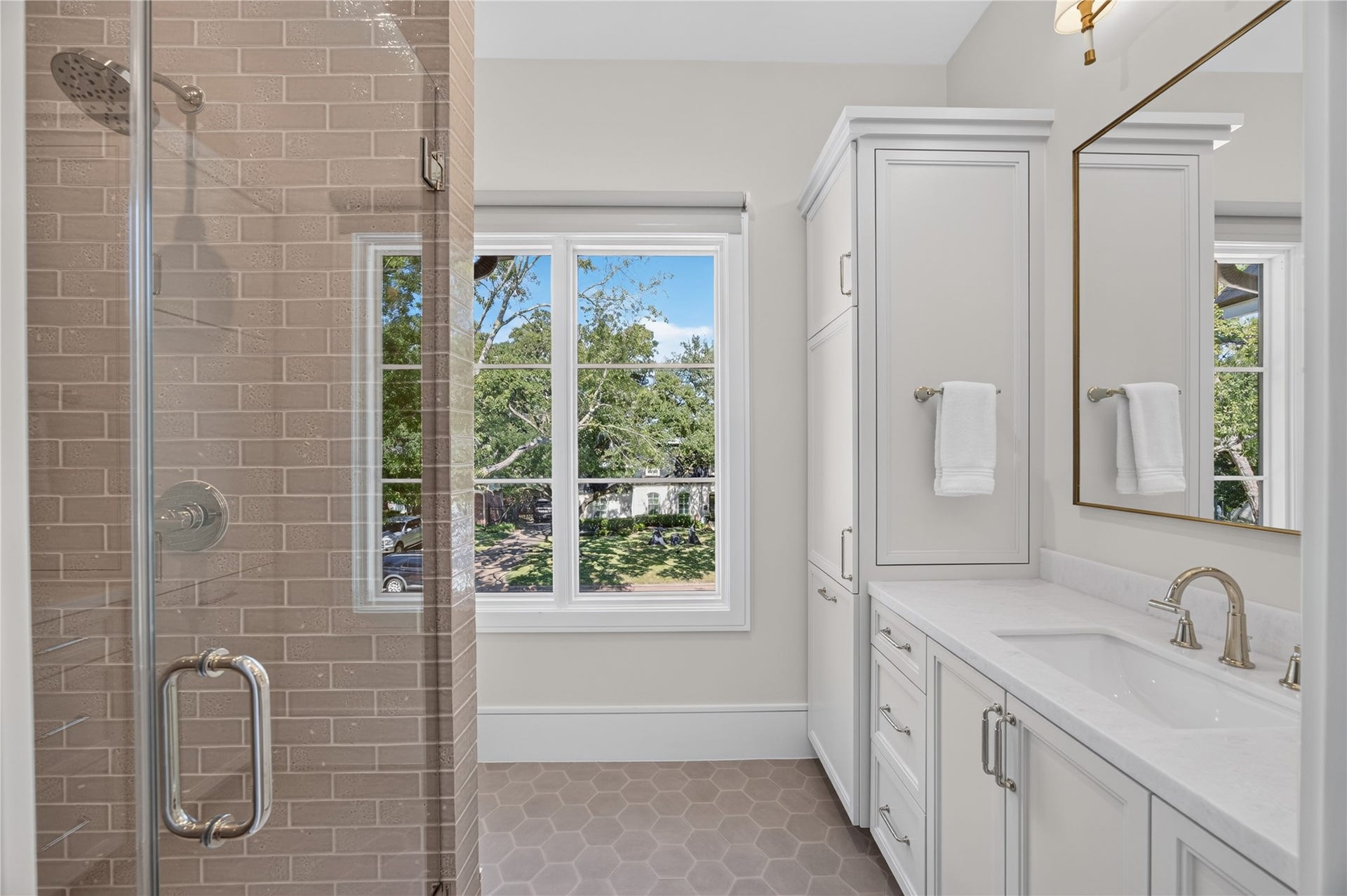 13915 Queensbury Lane, Houston, TX 77079 Beautifully appointed bathroom featuring a glass-enclosed shower with neutral tile surround, custom vanity with stone countertop, and additional built-in cabinetry. Natural light pours through the large window, complementing the stylish hexagonal tile flooring and refined finishes.