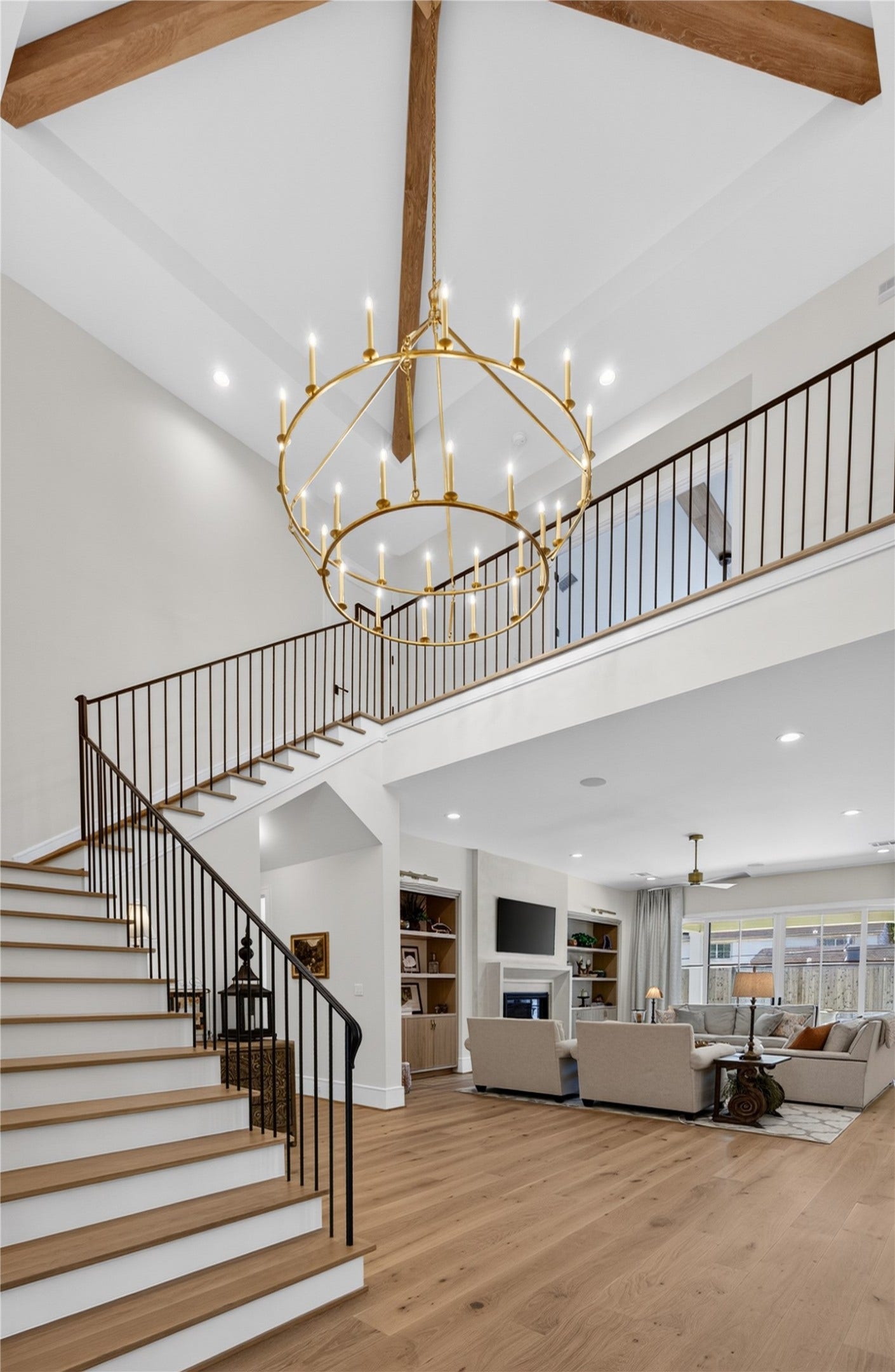 13915 Queensbury Lane, Houston, TX 77079 An impressive architectural centerpiece featuring soaring 25-foot ceilings, exposed wood beams, and a dramatic two-tier gold chandelier. The elegant staircase with wood treads and sleek black iron railings overlooks the spacious living area, creating a stunning sense of openness while showcasing the home's exceptional craftsmanship and thoughtful design.