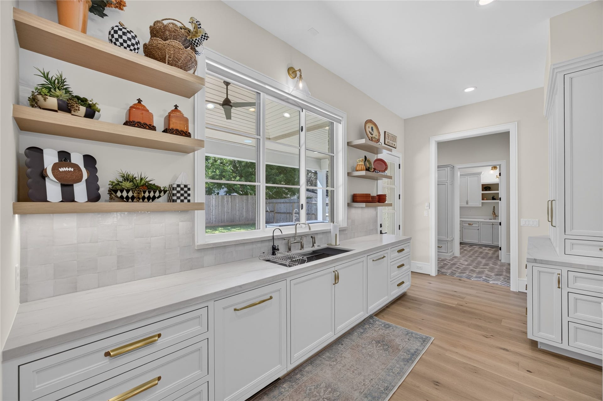 13915 Queensbury Lane, Houston, TX 77079 An alternate view of the kitchen highlights the expansive countertops, farmhouse-style sink beneath a large window overlooking the backyard, and beautiful floating wood shelves. Custom cabinetry, designer tile backsplash, and warm gold accents create a sophisticated yet inviting space.