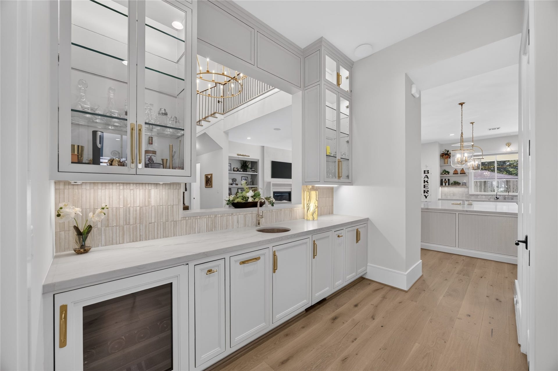 13915 Queensbury Lane, Houston, TX 77079 Beautifully designed wet bar featuring custom floor-to-ceiling cabinetry, glass-front display cabinets, elegant gold hardware, and a stylish tile backsplash. Complete with a built-in wine refrigerator, prep sink, and generous counter space, this thoughtfully designed area is ideal for entertaining and conveniently connects to the kitchen.