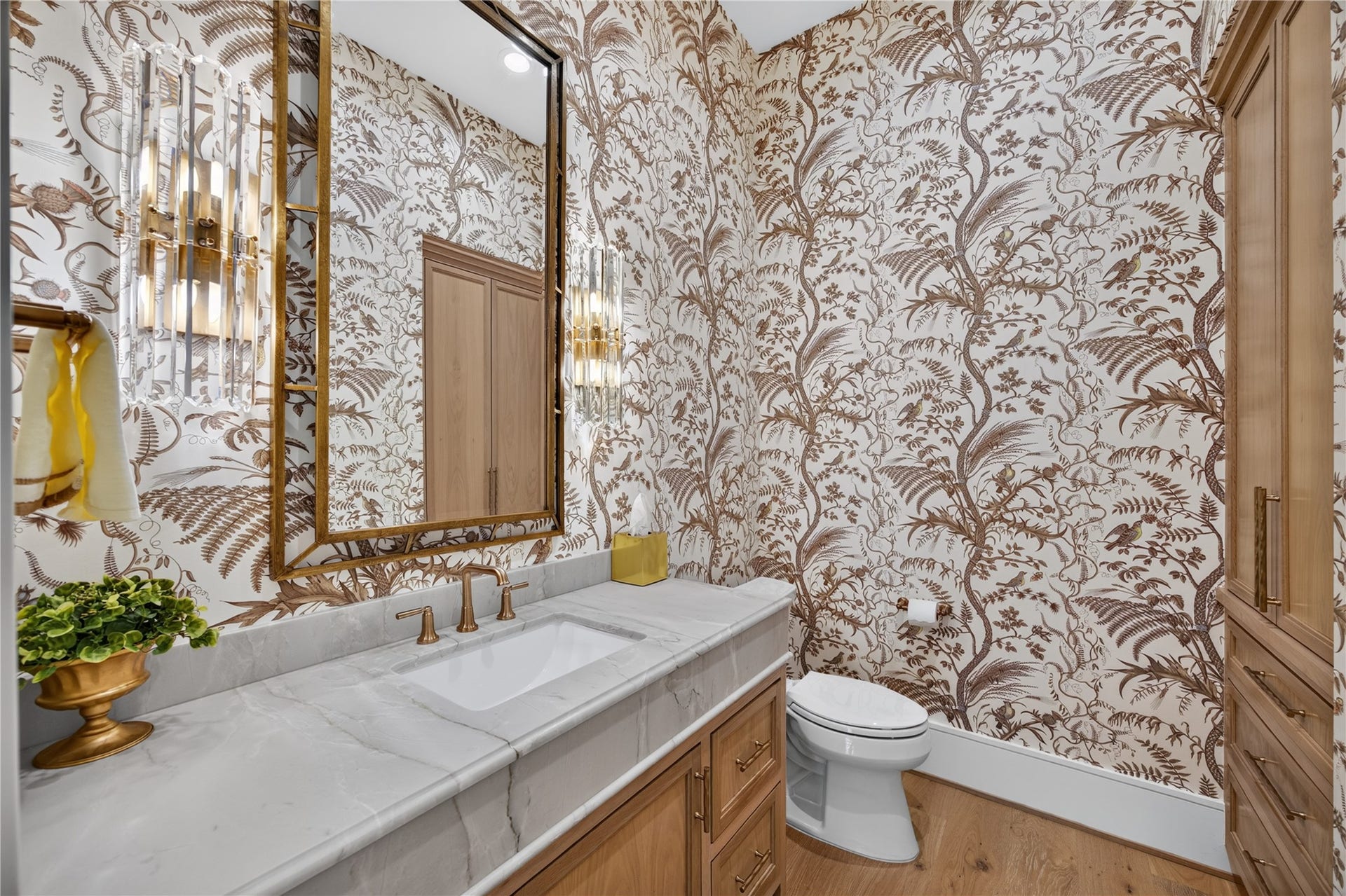 13915 Queensbury Lane, Houston, TX 77079 A stunning powder room that makes a statement with intricate botanical wallpaper, a beautiful stone countertop, custom natural wood cabinetry, and elegant brass fixtures. A gold-framed mirror and decorative wall sconces complete this sophisticated space, offering a memorable touch of luxury for guests.