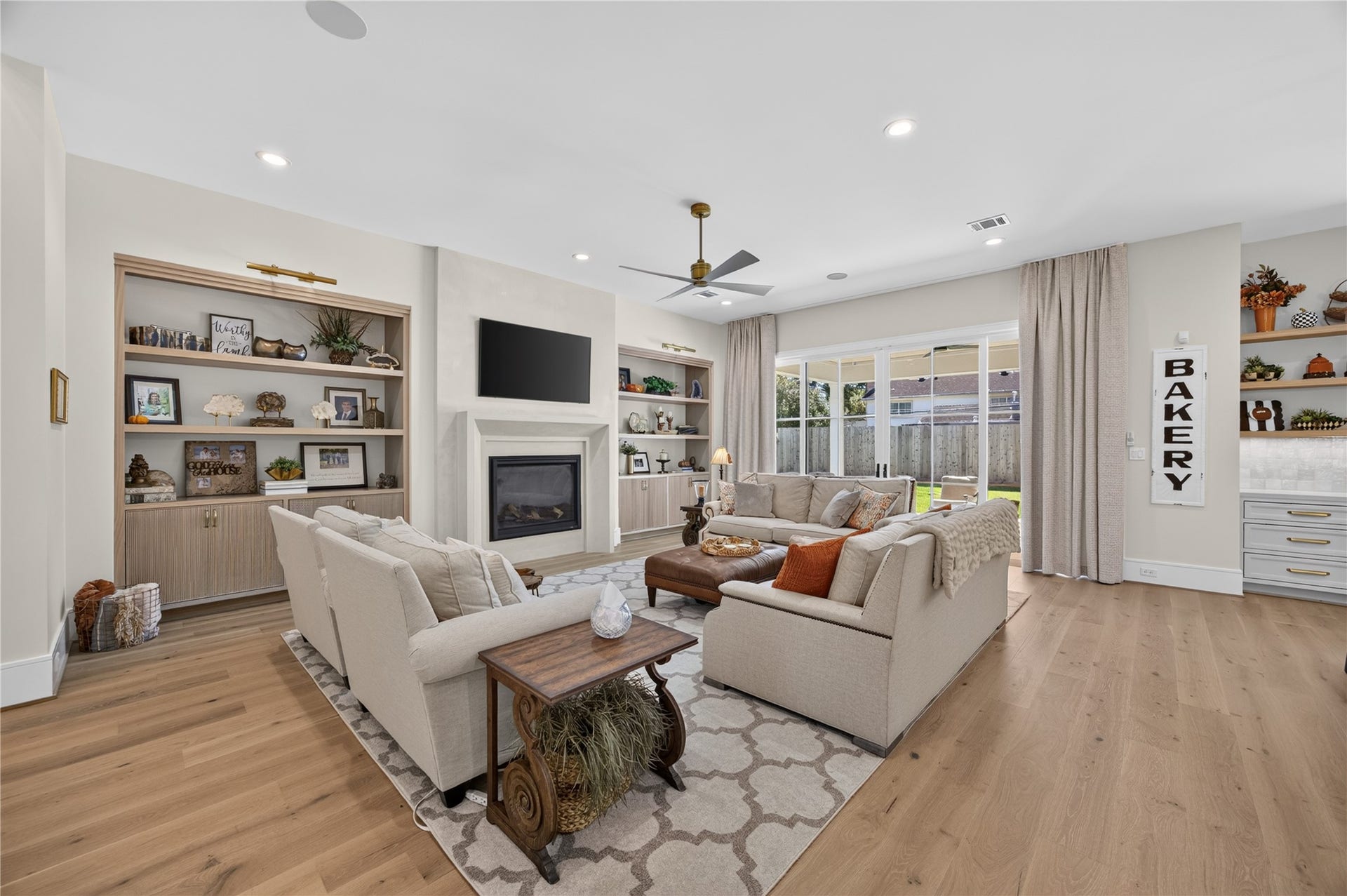 13915 Queensbury Lane, Houston, TX 77079 Beautifully appointed living room featuring wide-plank wood flooring, custom built-in shelving with lower cabinetry, and a decorative mock fireplace with a sleek surround. Expansive sliding glass doors bring in abundant natural light and provide seamless access to the covered outdoor patio, creating an inviting space for relaxing and entertaining.