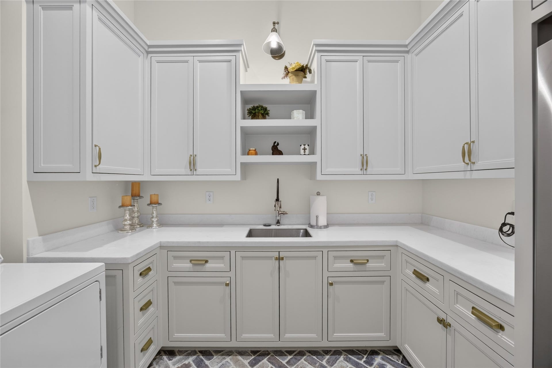 13915 Queensbury Lane, Houston, TX 77079 Spacious first-floor laundry room featuring extensive custom cabinetry, generous quartz-style countertops, a utility sink, and elegant gold hardware. Open shelving, designer lighting, and distinctive herringbone brick flooring combine functionality with sophisticated finishes, offering exceptional storage and workspace for everyday household needs.
