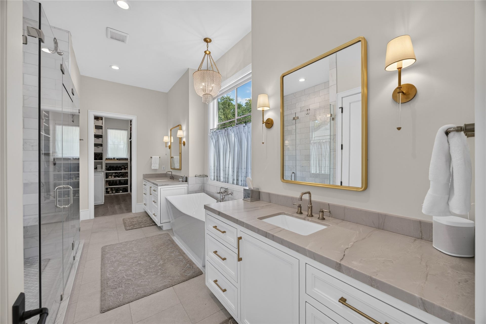13915 Queensbury Lane, Houston, TX 77079 Beautifully appointed primary bathroom featuring separate vanities with stone countertops, custom cabinetry, elegant gold accents, and designer lighting. A freestanding soaking tub, spacious glass-enclosed shower, and direct access to the oversized walk-in closet complete this luxurious retreat.