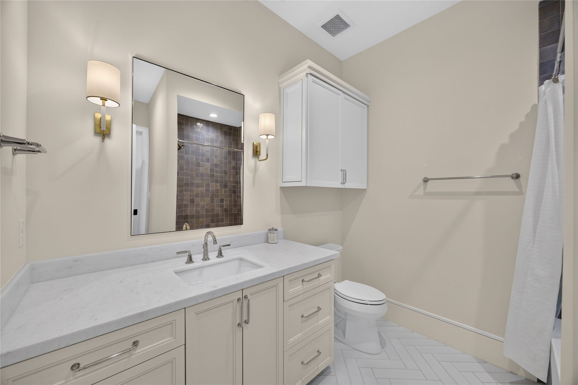 13915 Queensbury Lane, Houston, TX 77079 Beautifully finished secondary bathroom featuring an extended vanity with stone countertops, custom cabinetry, elegant wall sconces, and a large framed mirror. Herringbone tile flooring and a tiled tub/shower surround complete this thoughtfully designed space.