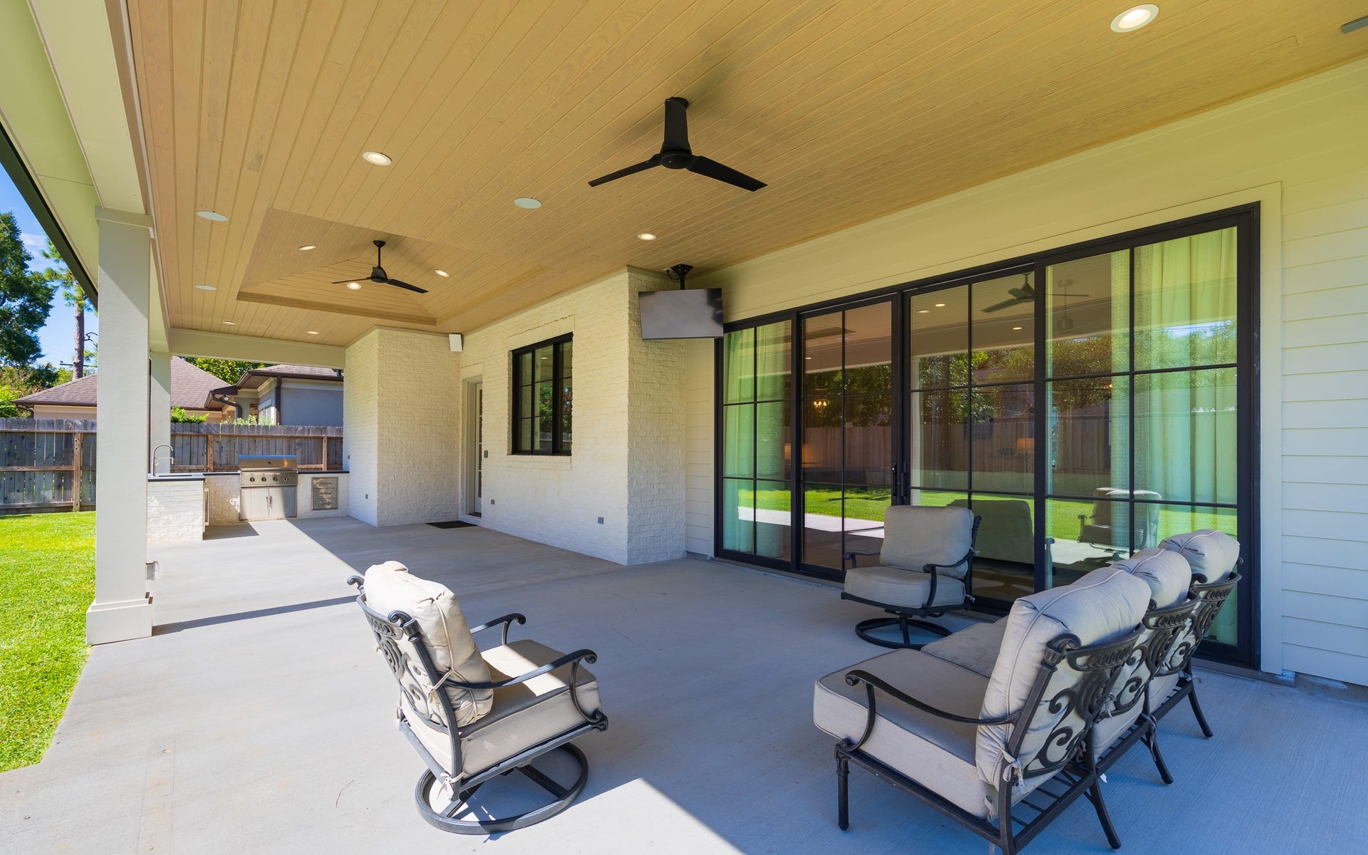 13915 Queensbury Lane, Houston, TX 77079 Seamlessly extending the home's living space outdoors, this spacious covered patio features beautiful wood-paneled ceilings, recessed lighting, ceiling fans, a mounted TV, and a fully equipped outdoor kitchen with built-in grill and sink. Expansive sliding glass doors connect the patio to the interior, creating an inviting space for year-round entertaining.