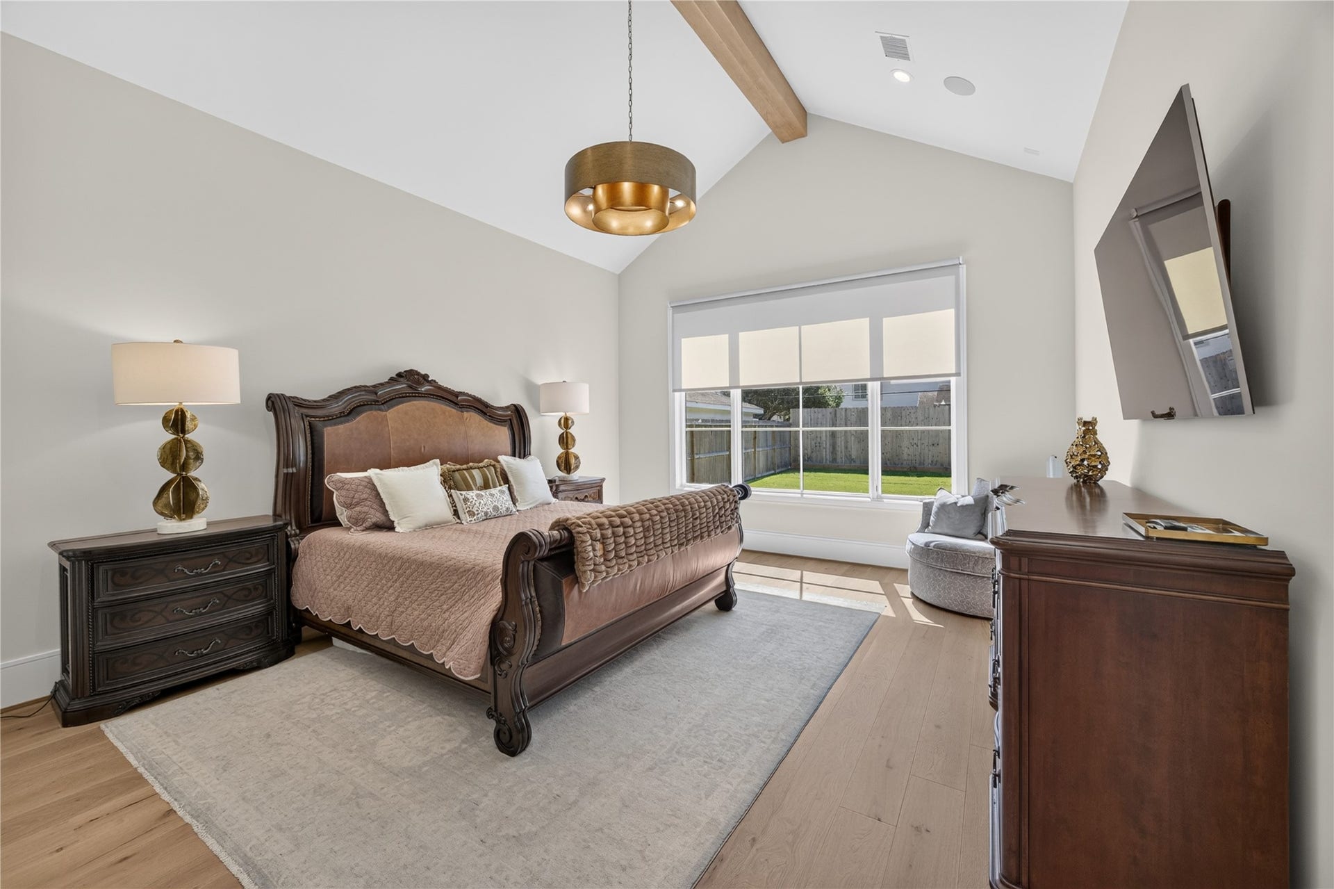 13915 Queensbury Lane, Houston, TX 77079 Spacious primary retreat featuring a vaulted ceiling with an exposed wood beam, elegant statement chandelier, and beautiful wide-plank wood flooring. An oversized window overlooking the backyard fills the room with natural light, creating a bright and relaxing atmosphere.