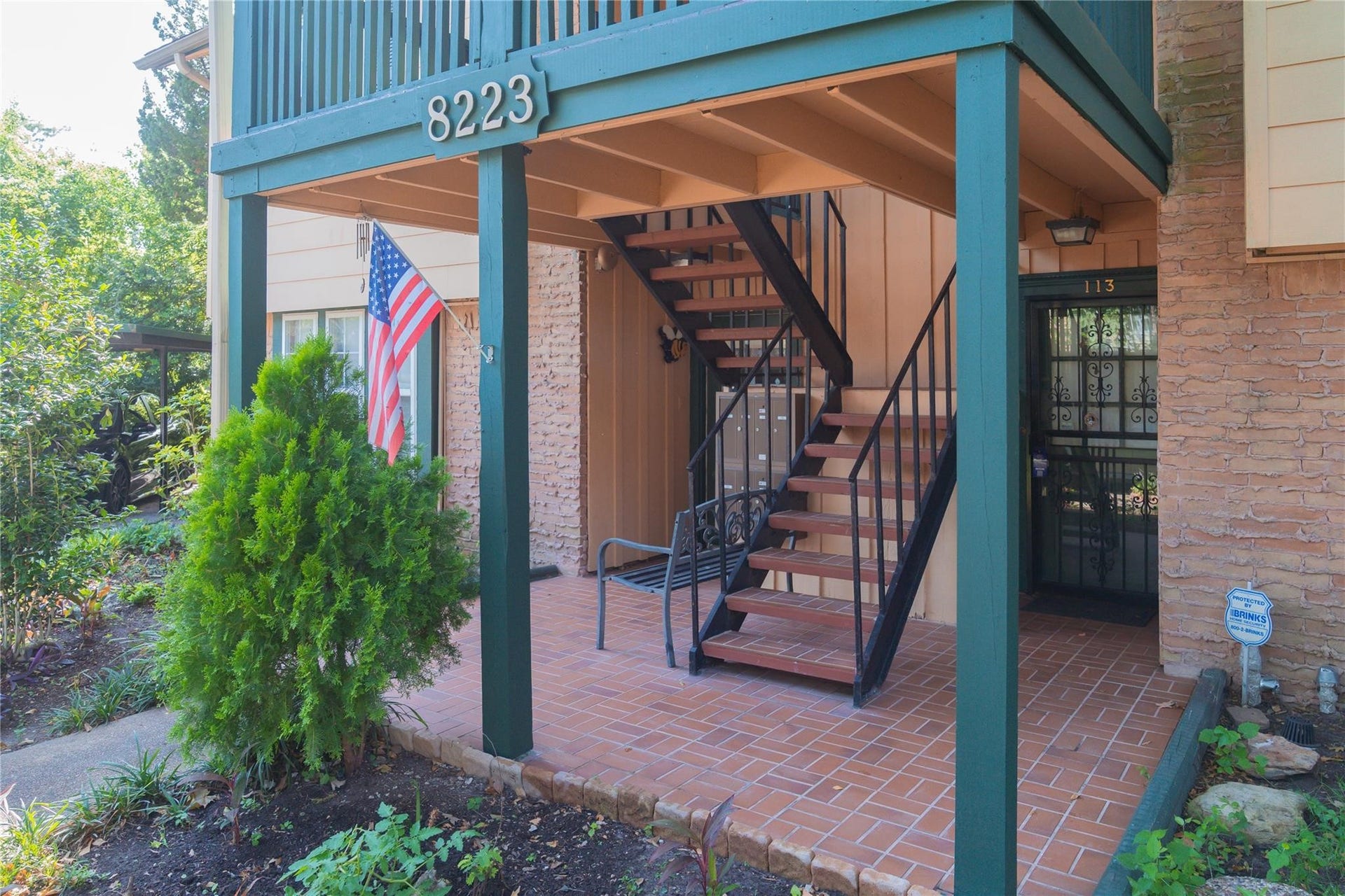 8223 Kingsbrook Road, #212, Houston, TX 77024 Charming entrance with a covered patio area, adorned with greenery and an American flag. Features a staircase leading to the upper level, brick flooring, and a secure gated door. Ideal for a welcoming, cozy feel.