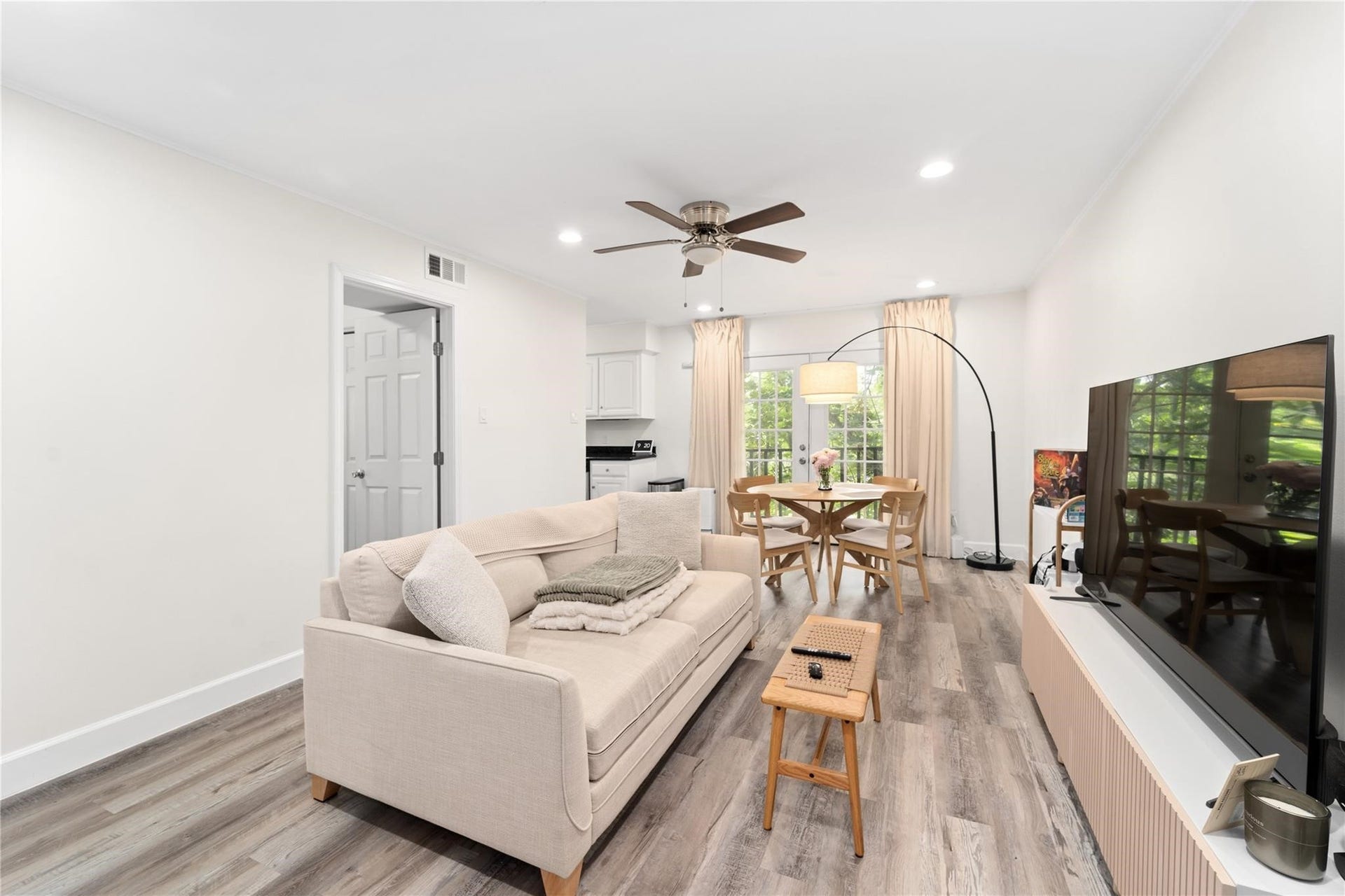 8223 Kingsbrook Road, #212, Houston, TX 77024 This bright, open-concept living area features modern flooring, a cozy sofa, and a large TV. The space flows into a dining area with natural light from large windows and glass doors, complemented by a ceiling fan and contemporary lighting.