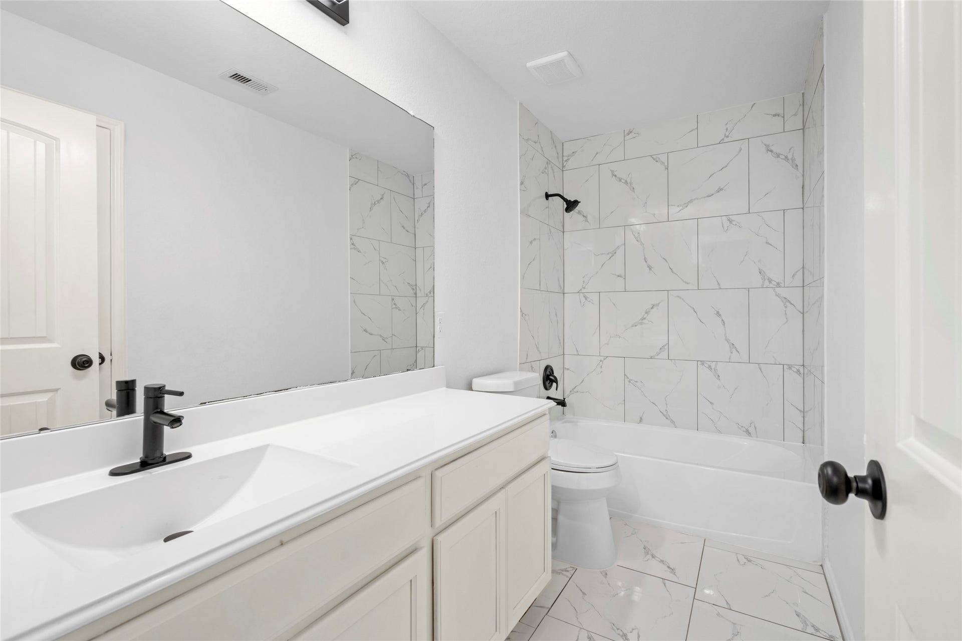 9423 Mineral Rock Lane, Richmond, TX 77407 Elegant bathroom featuring a sleek vanity with a modern black faucet, pristine white cabinetry, and stylish marble-patterned tiles surrounding the bathtub and flooring.