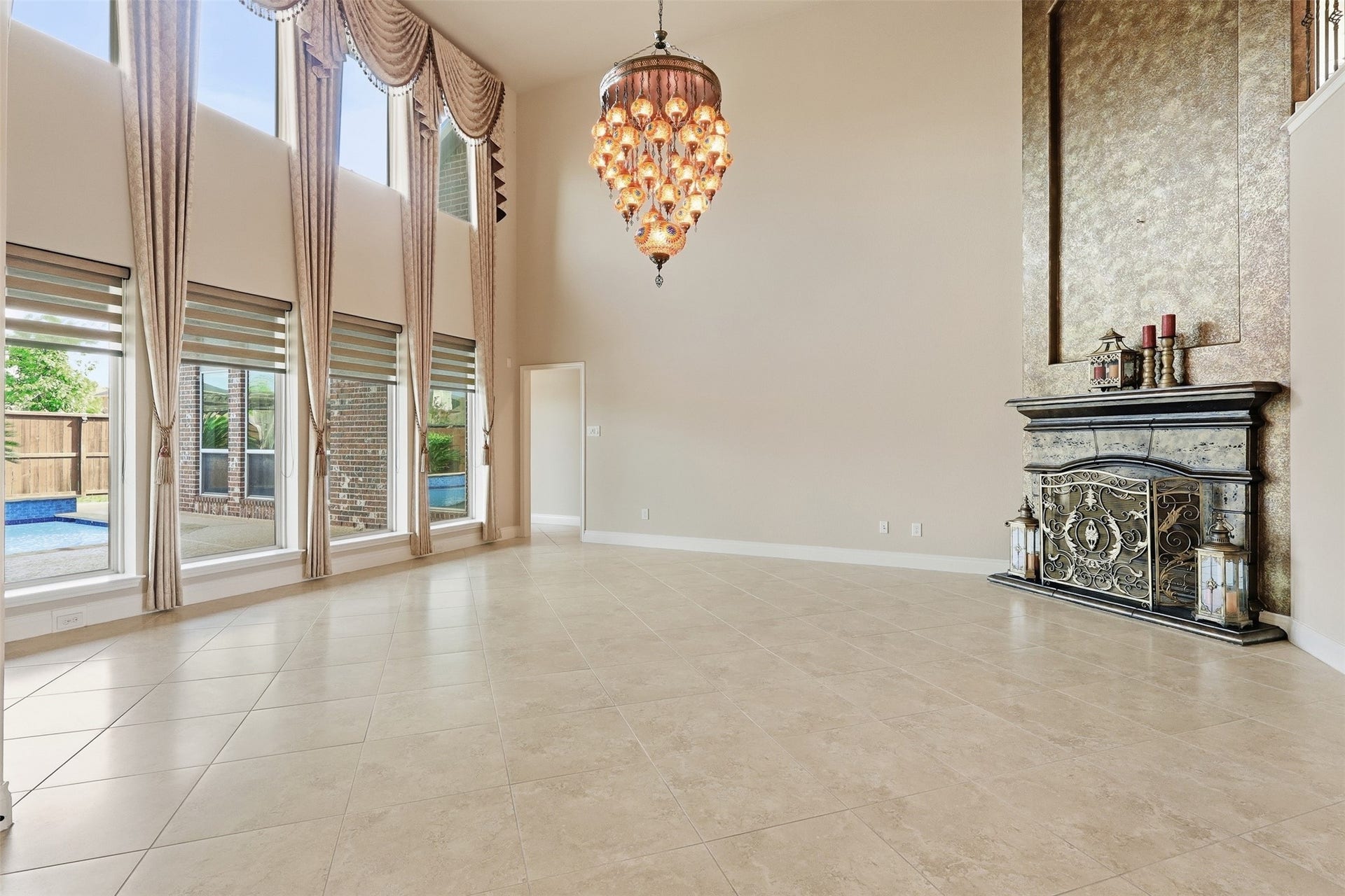4130 Clover Ridge Lane, Sugar Land, TX 77479 This elegant living space features a high ceiling with a grand chandelier, large windows with drapes offering abundant natural light and a view of the outdoor pool. The ornate fireplace and neutral tile flooring add a touch of luxury.