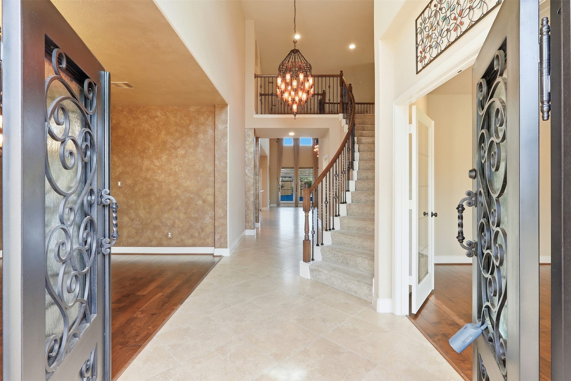 4130 Clover Ridge Lane, Sugar Land, TX 77479 This elegant entryway features ornate wrought iron doors, a grand staircase with carpeted steps and wooden railings, and a stunning chandelier, all set against a backdrop of warm-toned walls and tile flooring.