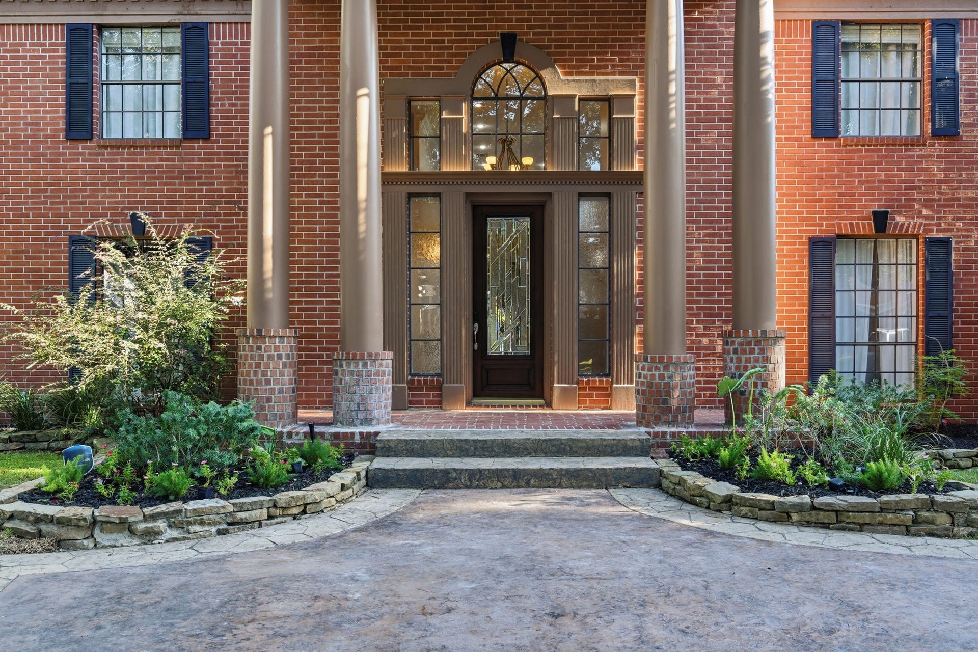 An impressive covered entrance makes a grand first impression, with towering columns framing the dramatic two-story entry. An elegant leaded-glass front door, timeless red brick exterior, & window shutters complement the home's stately architecture, creating a distinguished & welcoming arrival.