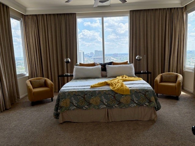 2016 Main Street, #PH 3, Houston, TX 77002 Spacious bedroom with panoramic city views, featuring a cozy bed adorned with a decorative throw, flanked by elegant armchairs and floor-length drapes.