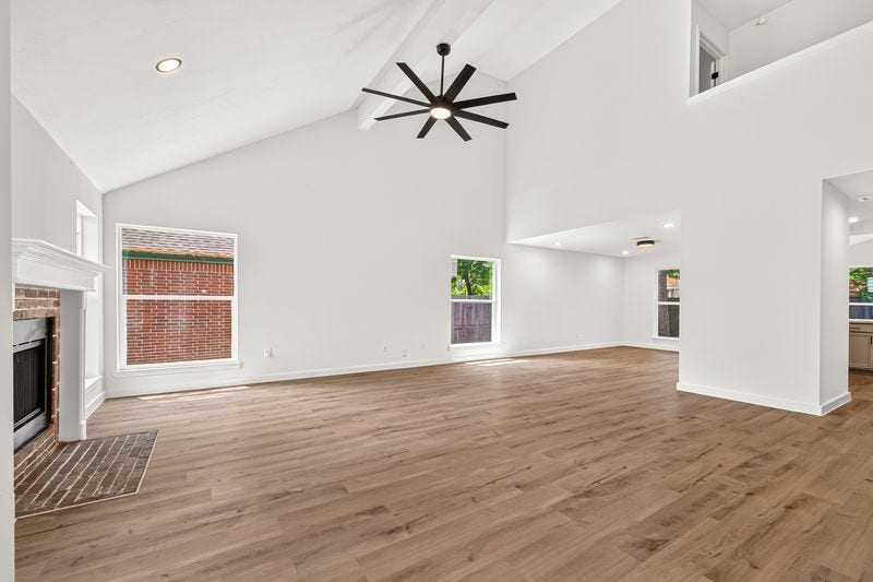 Spacious living area with vaulted ceilings, modern fan, and abundant natural light, featuring elegant wood flooring and a cozy brick fireplace.