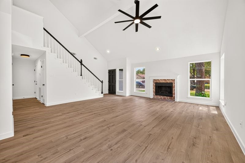 Spacious living area with vaulted ceilings, a modern ceiling fan, and abundant natural light, featuring a charming brick fireplace and elegant wood flooring.