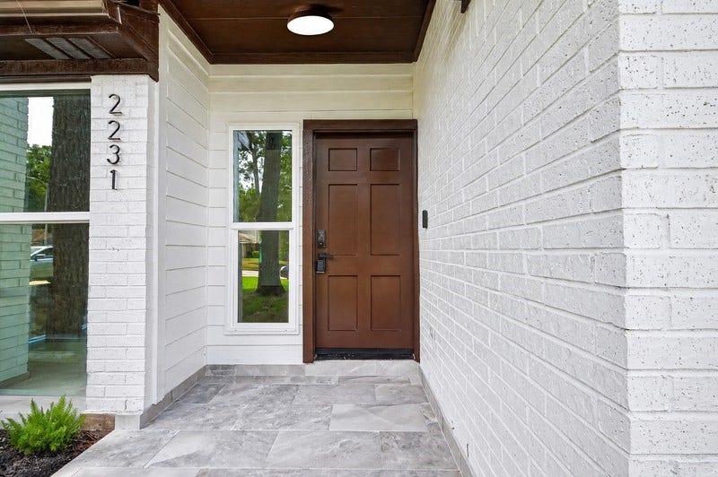 Charming entryway featuring a rich wooden door, elegant tile flooring, and a bright window, set against a crisp white brick facade.
