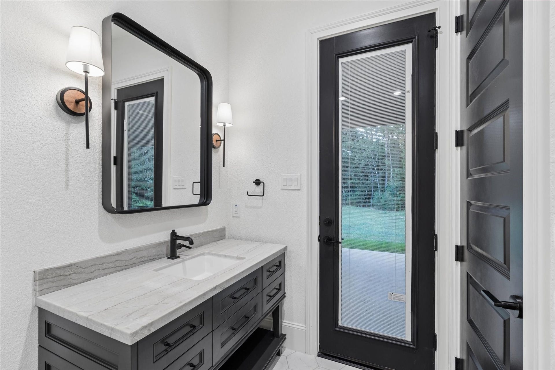 This beautifully appointed pool bathroom continues the homes sophisticated design with a custom vanity, stone countertop, oversized framed mirror, and coordinated black fixtures. A glass-paneled exterior door provides direct access to the covered patio and backyard, allowing guests to enter without moving through the main living areas. Designer sconces, elevated millwork, and timeless finishes create a polished and functional space perfectly.