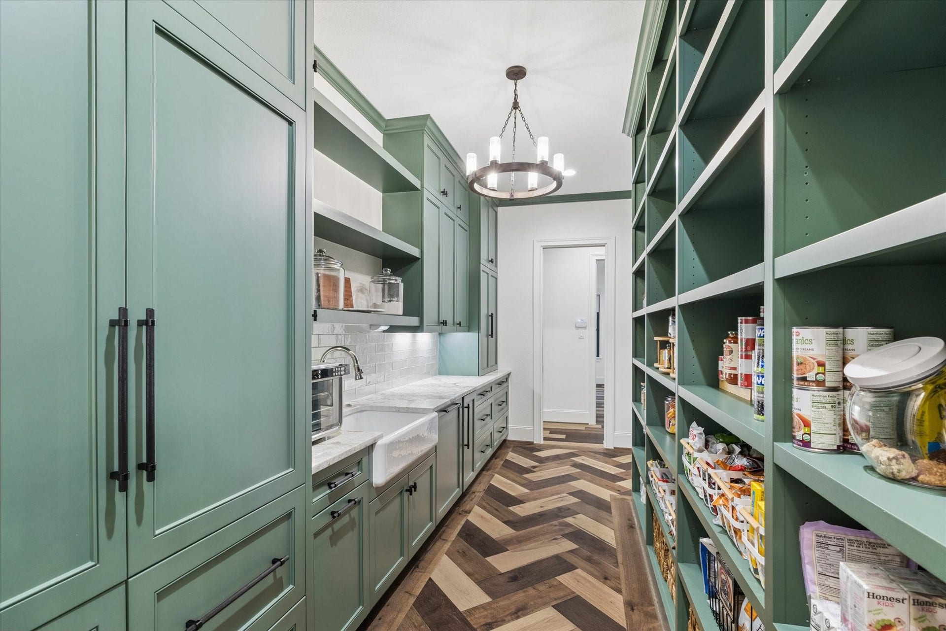 This additional view showcases the exceptional storage capacity and designer craftsmanship that define the prep pantry and scullery. Floor-to-ceiling cabinetry, expansive open shelving, pullout organization systems, and generous countertops ensure every item has its place. Rich green cabinetry, a handcrafted-inspired backsplash, farmhouse sink, designer lighting, and herringbone hardwood flooring create a sophisticated secondary kitchen that seamlessly complements the homes luxurious main culinary space.