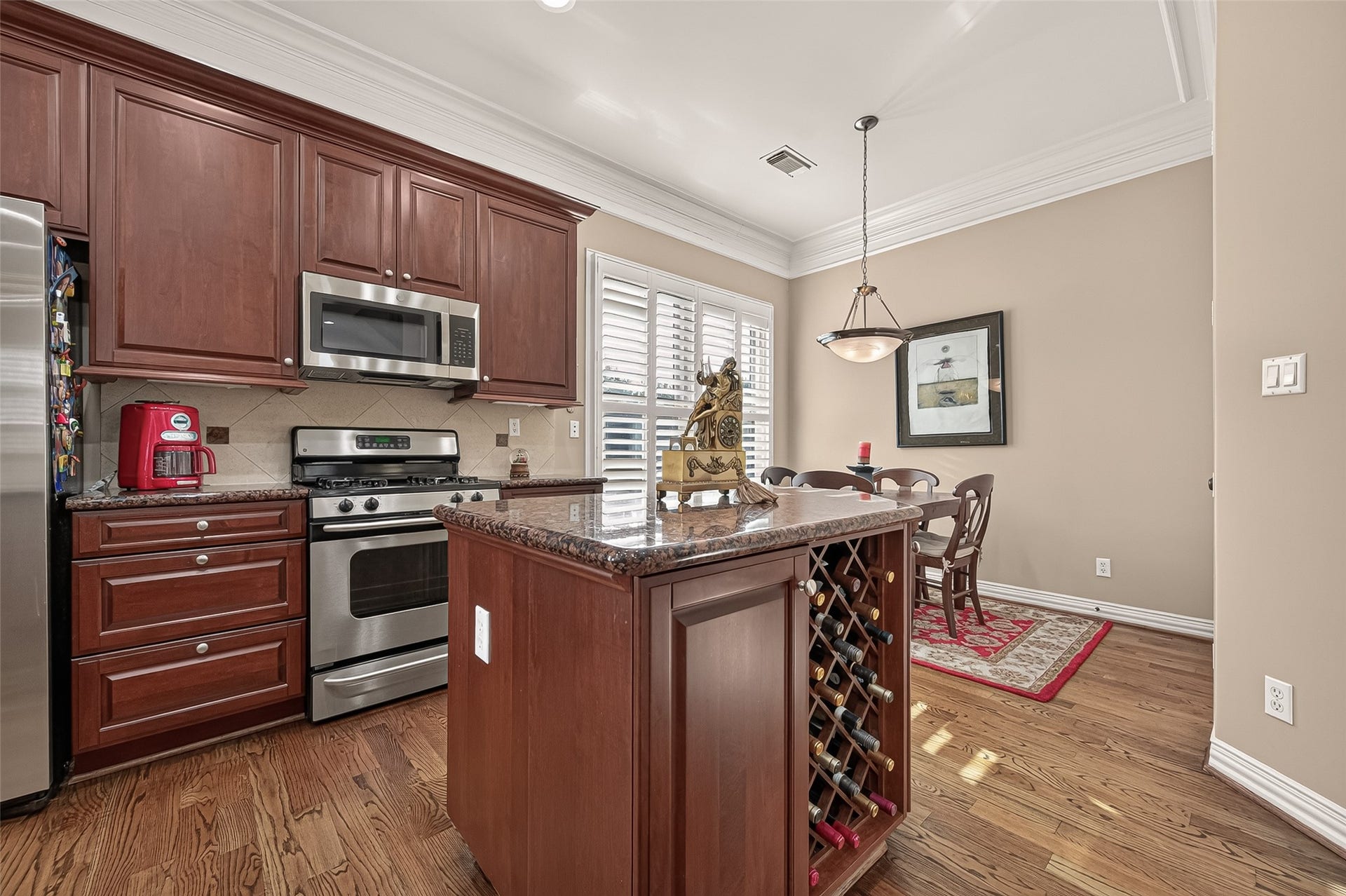 4003 Bellefontaine Street, Houston, TX 77025 Inviting kitchen and breakfast nook featuring a granite-topped island with built-in wine storage, rich wood cabinetry, and a stainless steel gas range. Hardwood floors, crown molding, and plantation shutters create a warm setting for everyday dining.