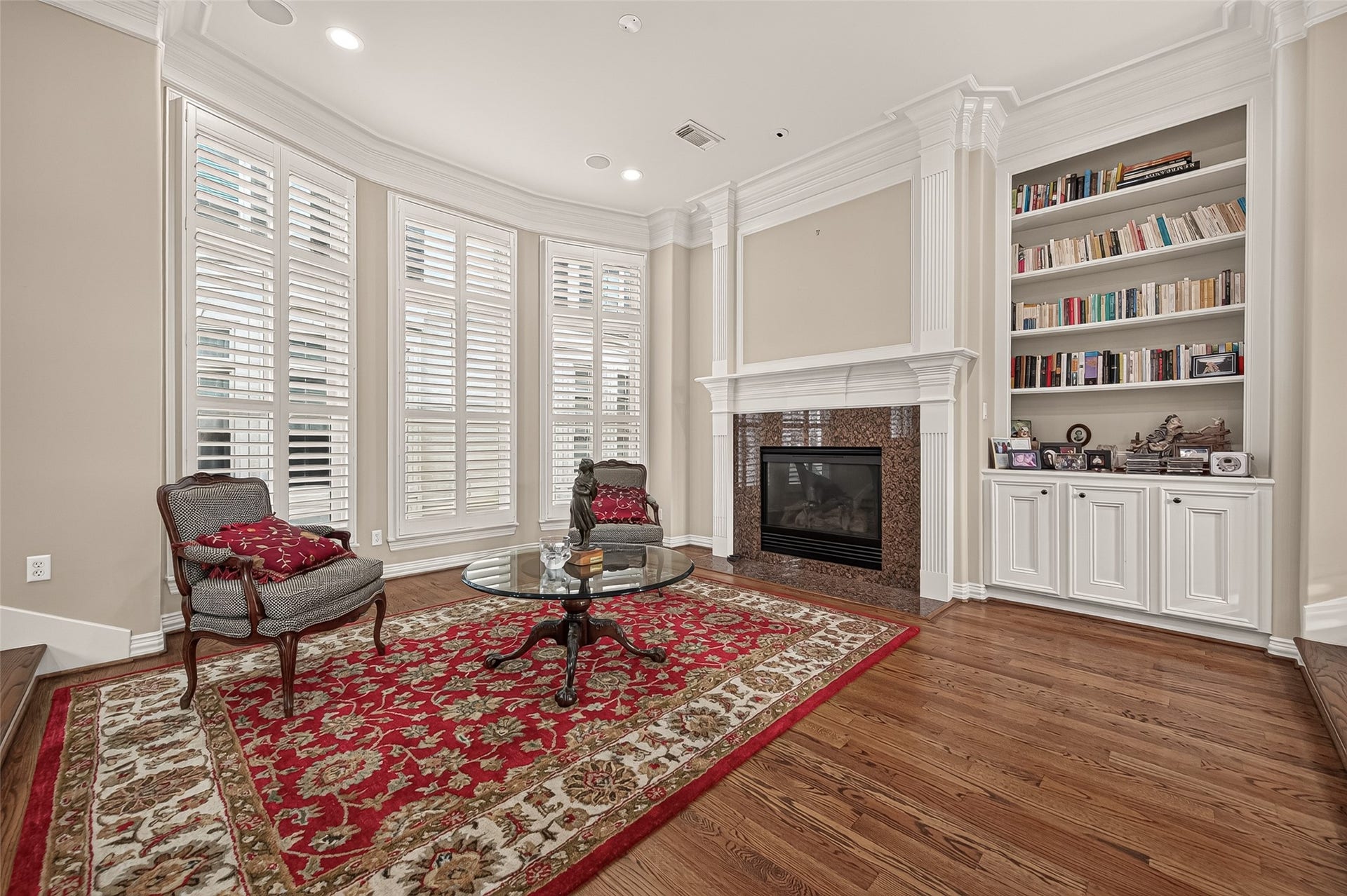 4003 Bellefontaine Street, Houston, TX 77025 An elegant, light-filled living room featuring rich hardwood floors, soaring windows with plantation shutters, and a fireplace framed by detailed millwork. Custom built-in shelving and cabinetry provide beautiful display space and generous storage.