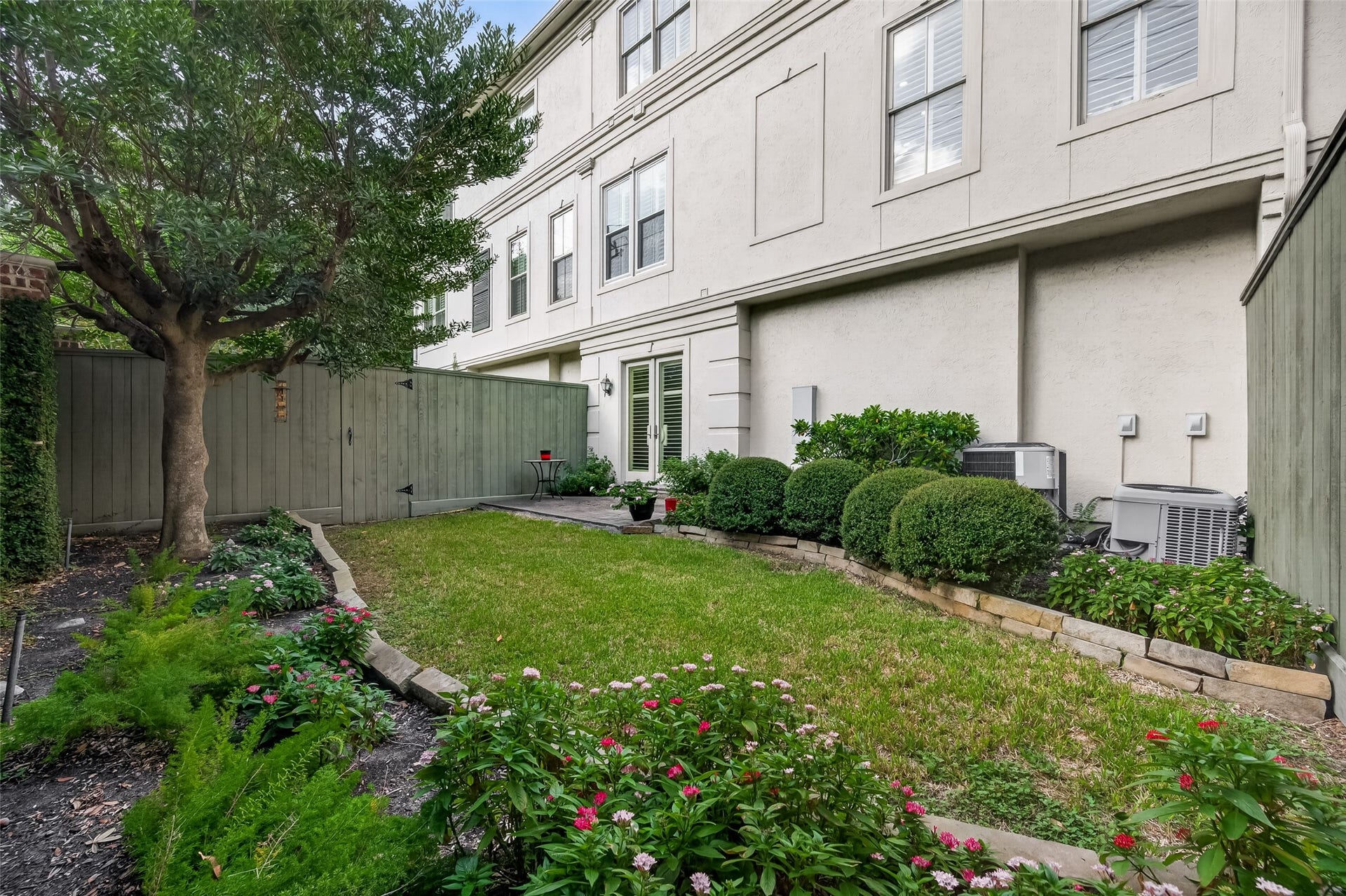 4003 Bellefontaine Street, Houston, TX 77025 Private garden retreat featuring a grassy lawn, mature shade tree, and stone-bordered beds filled with flowering plants and manicured shrubs. A paver patio connects to the first-floor bedroom or office through double glass doors, offering an inviting setting for outdoor relaxation.