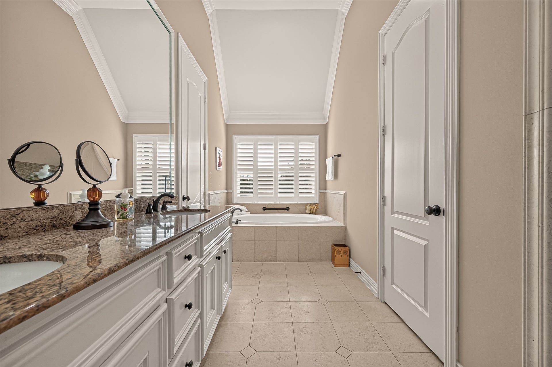 4003 Bellefontaine Street, Houston, TX 77025 Elegant primary bathroom featuring an extended granite vanity with dual sinks and abundant cabinetry, tile flooring, and a vaulted ceiling. A jetted soaking tub beneath a window with plantation shutters creates a relaxing retreat.
