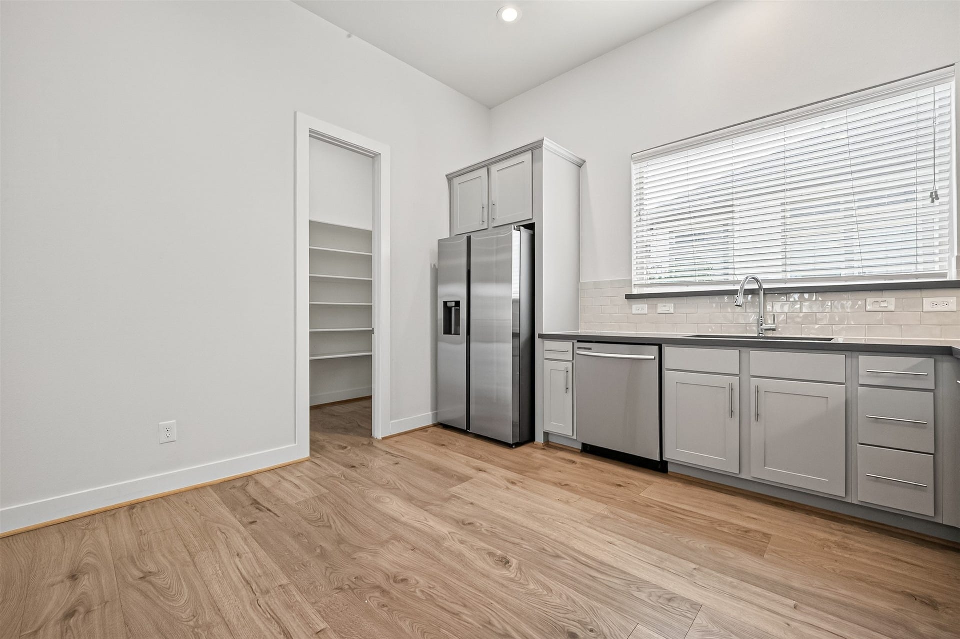 Modern kitchen with sleek stainless steel appliances, gray cabinetry, and a spacious pantry. Bright natural light fills the space through a large window, highlighting the elegant Swiss flooring.