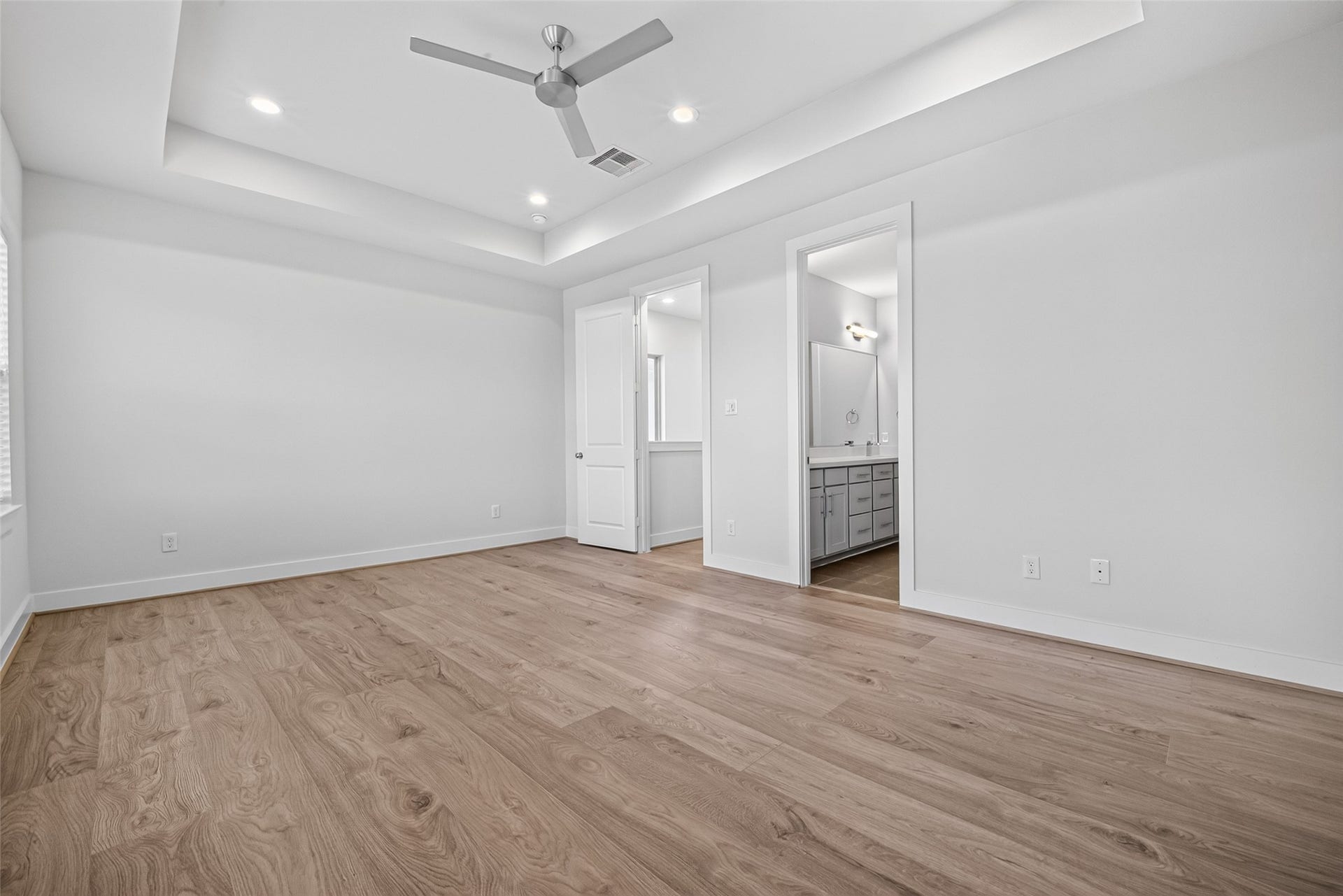 Spacious Master bedroom with elegant tray ceiling, modern ceiling fan, and warm wood LVP flooring, leading to an en-suite bathroom with contemporary fixtures.