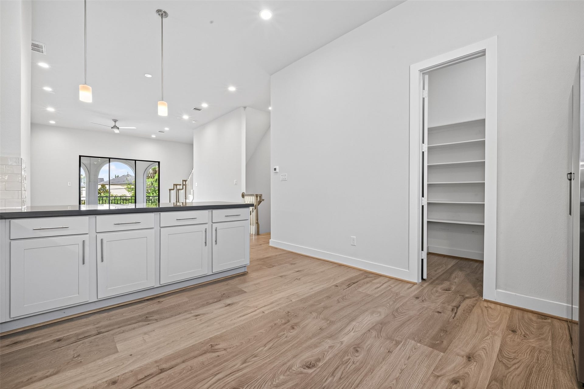 Modern kitchen with sleek white cabinetry and a spacious island, featuring elegant pendant lighting. The open layout flows into a bright living area with large sliding doors, while a convenient walk-in pantry offers ample storage space.