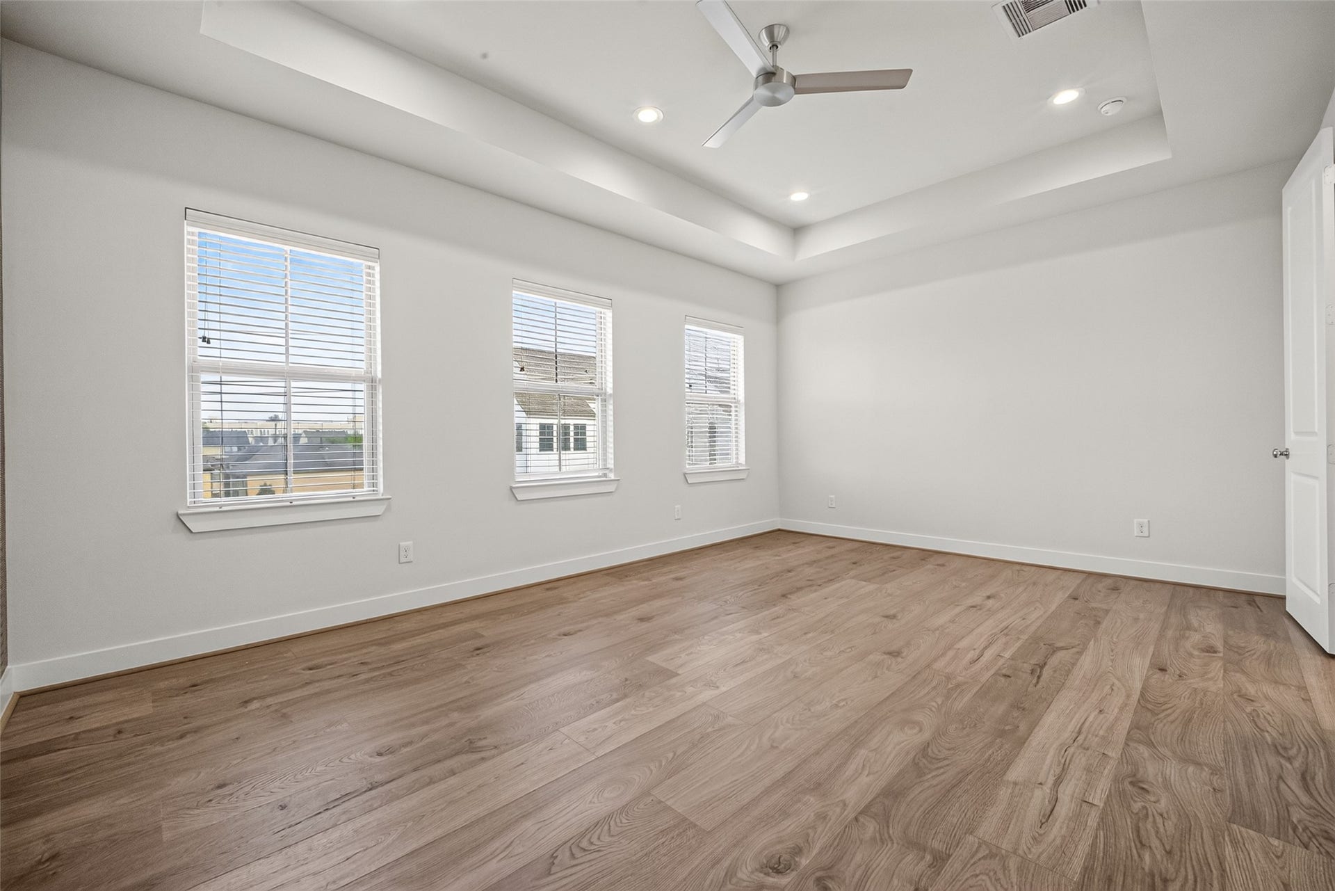 Spacious Master bedroom with elegant tray ceiling, abundant natural light from three large windows, and sleek Swiss floors.