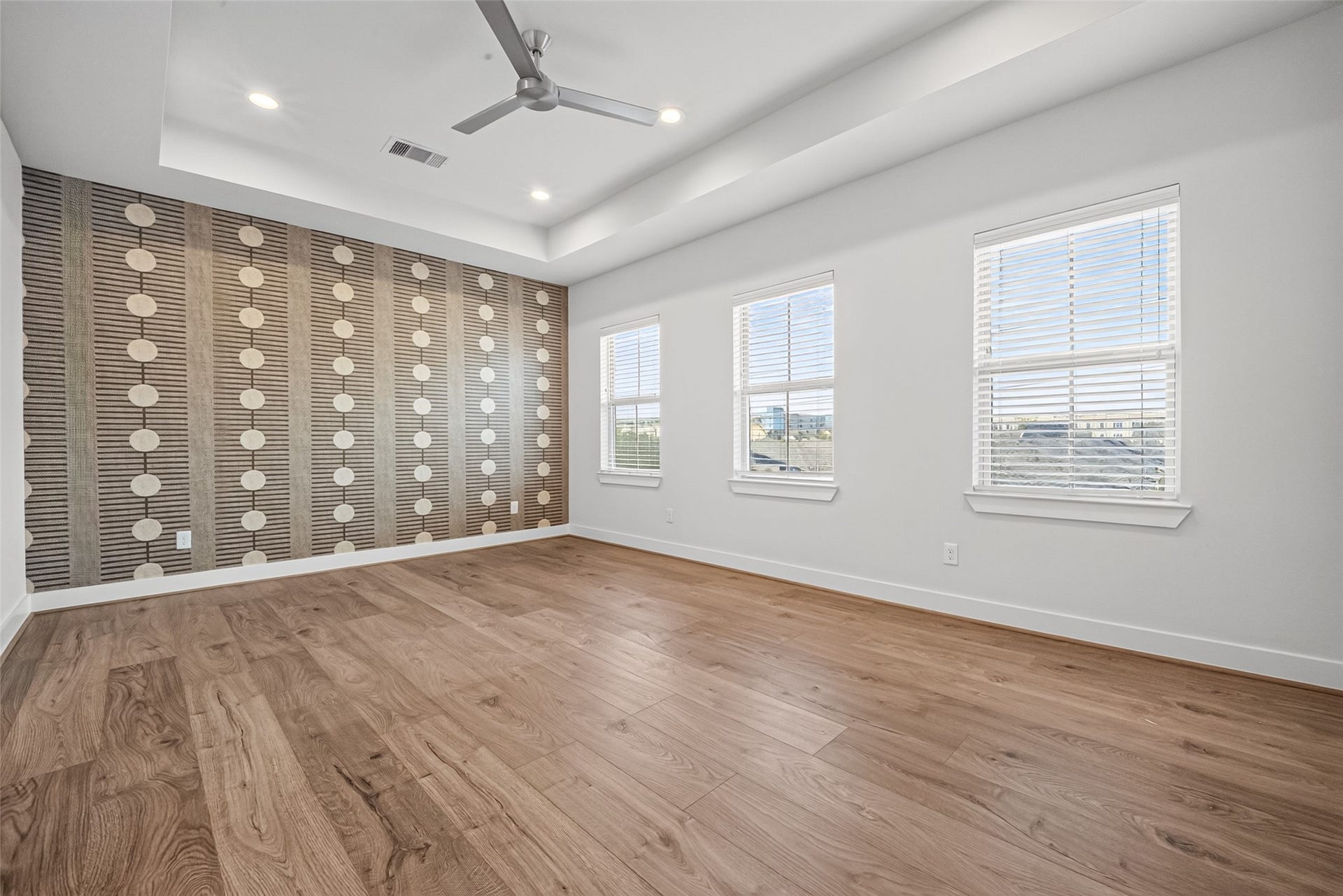 Spacious Master bedroom featuring elegant wood like flooring, a stylish accent wall, recessed lighting, and plenty of natural light through double windows.