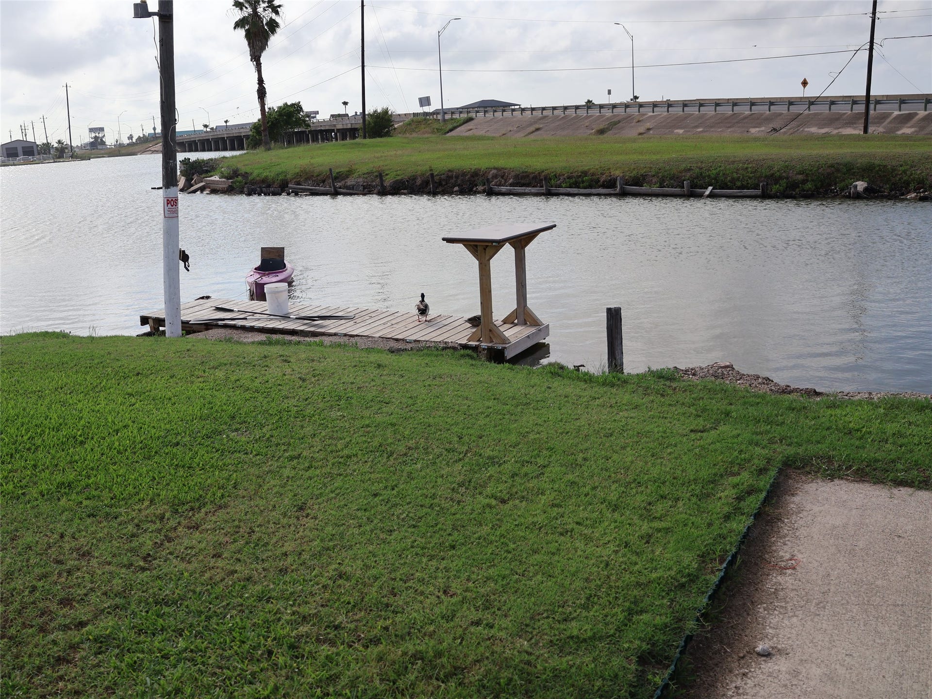 3401 Highway 6, Hitchcock, TX 77563 This photo shows a serene waterfront view with a small wooden dock on a grassy bank. The area offers a peaceful setting with open water and nearby road access. Ideal for enjoying outdoor activities and relaxation by the water.