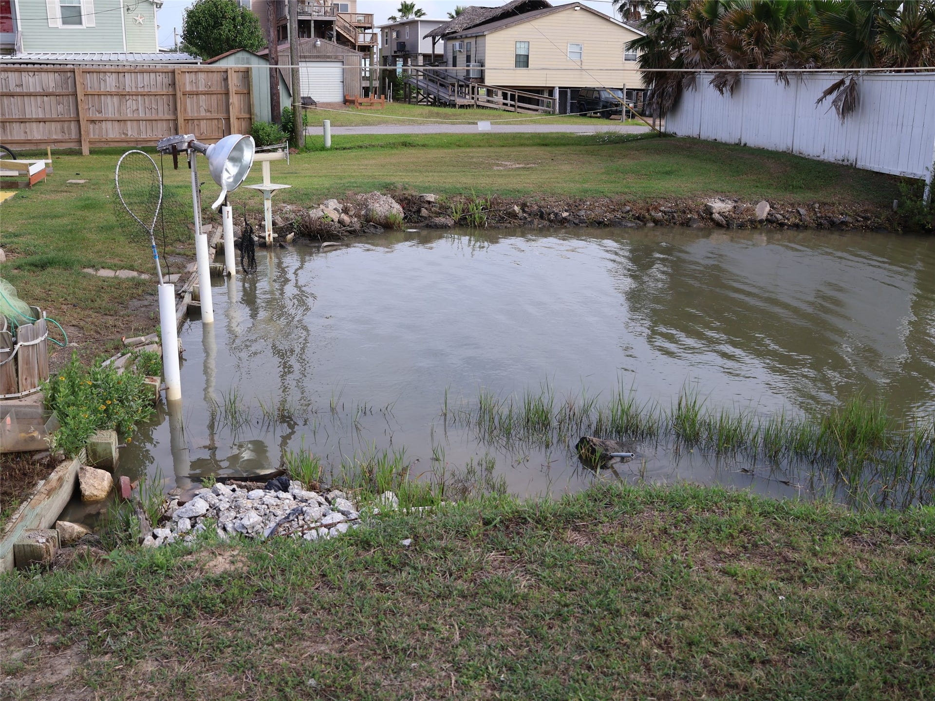 3401 Highway 6, Hitchcock, TX 77563 This photo shows a backyard with a small waterway, surrounded by grass and a wooden fence. There's a light fixture and a net nearby, allowing for fishing activities.