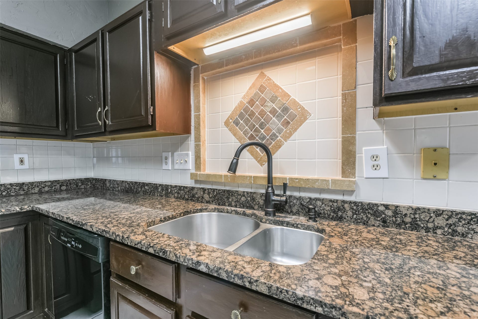 2509 S Mission Circle, Friendswood, TX 77546 Charming kitchen features dark wood cabinetry, granite countertops, and a stylish tile backsplash with a mosaic design above the dual sink, complemented by under-cabinet lighting.